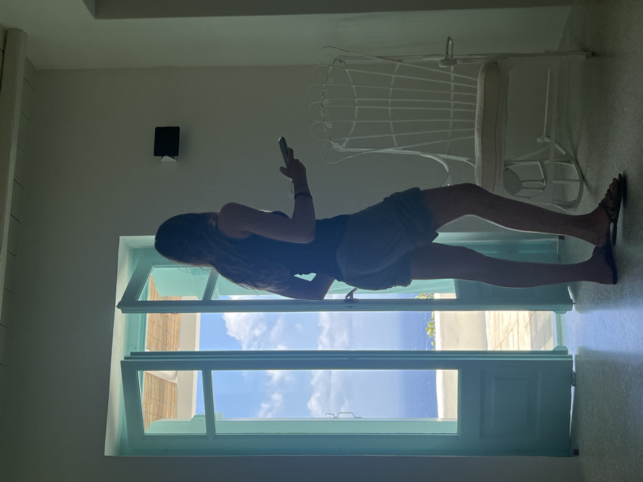 The image depicts the interior of a structure, likely a residence, with an open doorway leading to an exterior scene in Tourlos, Greece. An adult female, with long dark hair, stands in the foreground facing away from the camera, silhouetted against the bright light from the doorway. She wears a dark tank top and light-colored shorts, with patterned flat sandals. Her right hand holds a light-colored smartphone horizontally. To her right, a white, ornate metal rocking chair with a cream cushion is positioned. A black, rectangular device is mounted on the white wall above and to the left of the rocking chair. The ceiling has visible white wooden beams, and the floor is a light, possibly tiled or concrete, surface.
Through the open, light turquoise-colored double doors, a bright, clear blue sky with scattered white clouds is visible. Below the sky, a vast expanse of dark blue sea extends to the horizon. Immediately outside the doorway, a light-colored outdoor terrace or balcony is present, featuring a section of a light-colored, possibly straw, canopy above. Green foliage is discernible on the distant land near the coastline. The strong exterior light backlights the interior scene.