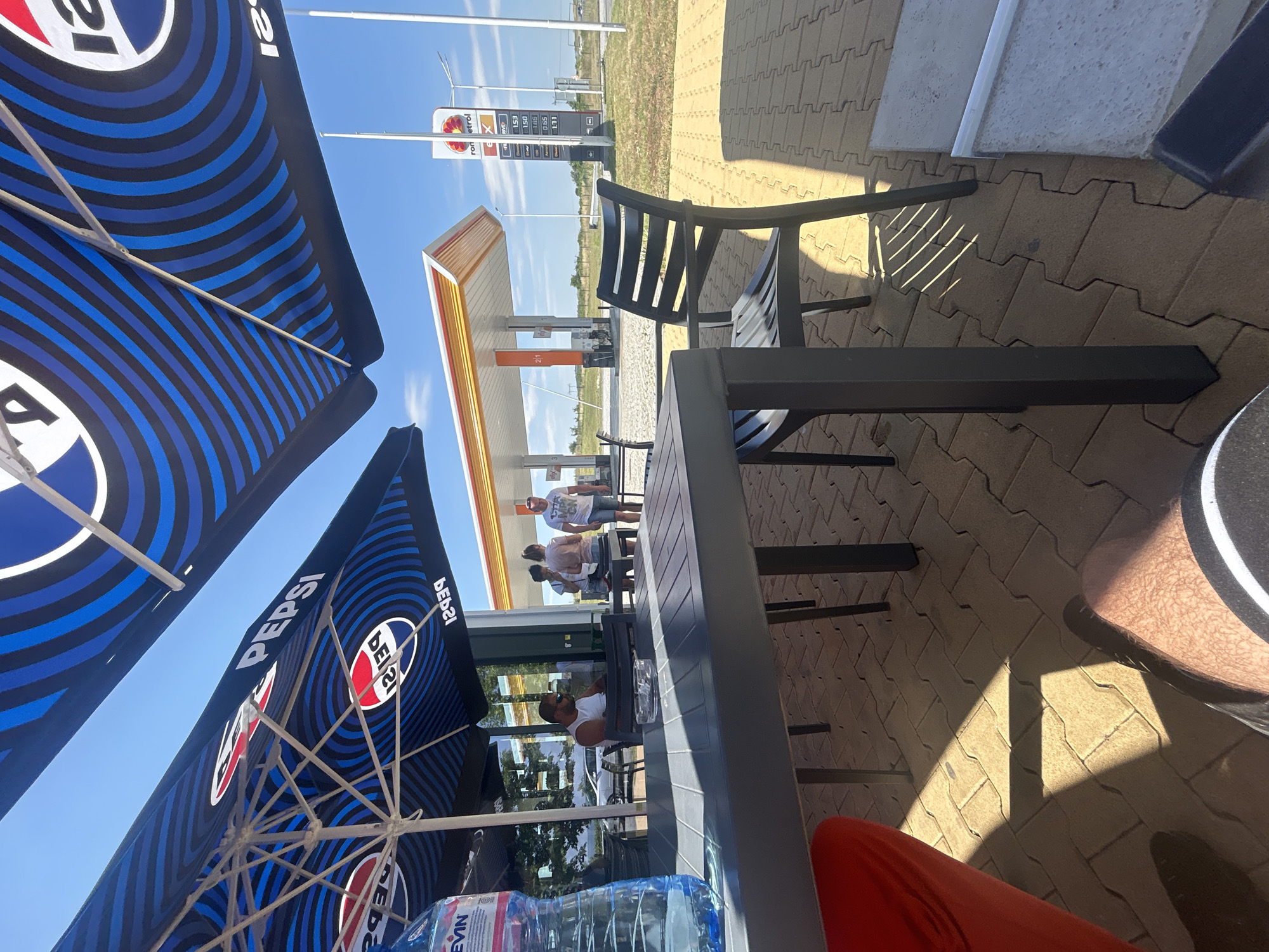 The image captures a sunny outdoor seating area with a gas station in the background. Two large umbrellas with the Pepsi logo provide shade over a table and chairs. On the table, a large bottle of water and an ashtray are visible. A man wearing a white tank top and sunglasses is seated at the table, facing left. In the background, two women and another man are standing near a building. The gas station has a distinctive orange and white canopy, and a sign displays fuel prices. The ground is paved with light brown interlocking bricks. The sky is clear and blue. The scene appears to be a rest stop or service area on a road, possibly near Polsko Padarevo, Bulgaria, given the presence of the "rom petrol" sign which indicates a Bulgarian petrol station chain.