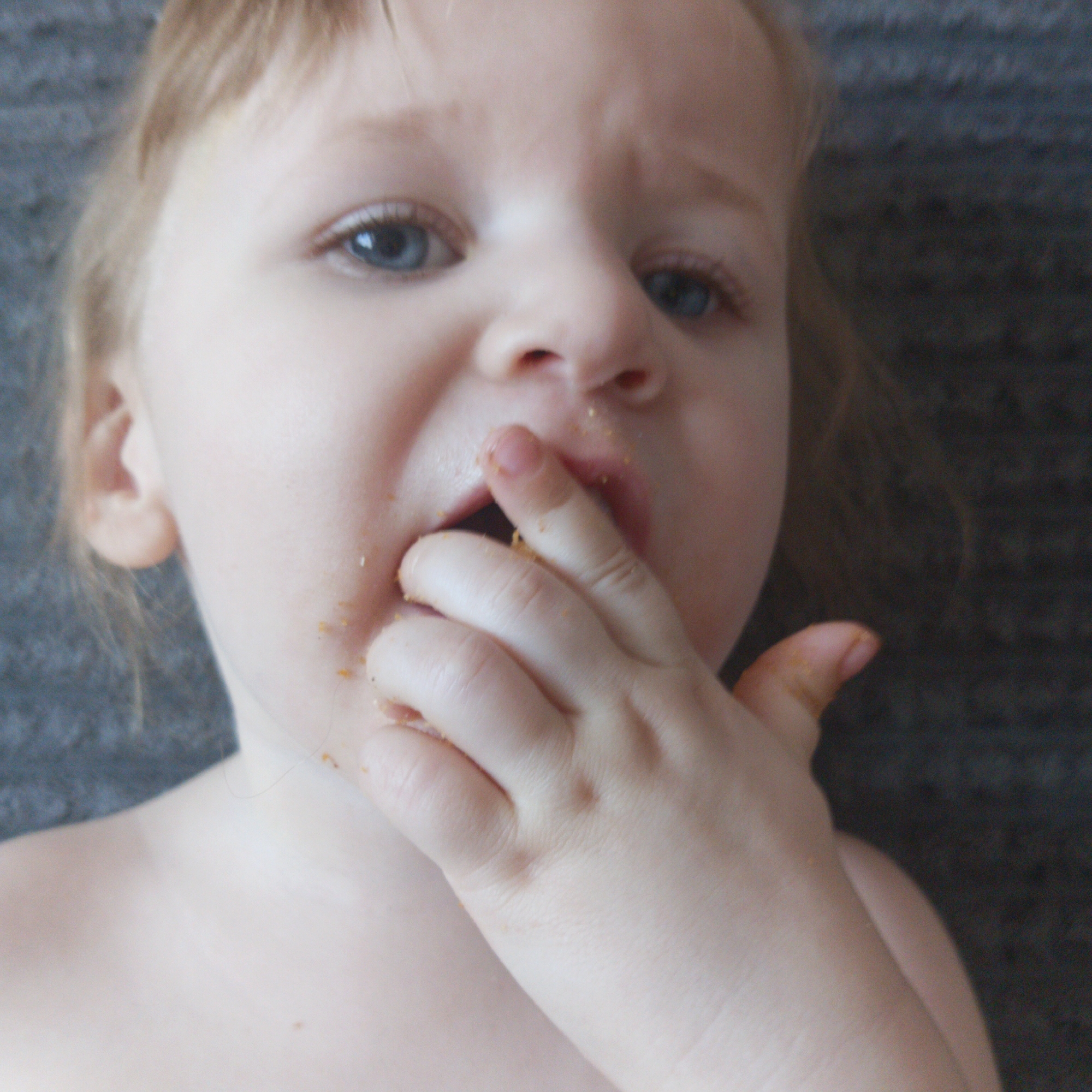 A close-up, candid portrait features a young child, likely a toddler, actively engaged in eating. The child has fair skin, light-colored hair, and striking blue eyes that look directly towards the viewer. Their mouth is slightly open, revealing the process of eating.
The child's right hand is brought up to their face, with fingers either in or very close to their mouth. The fingers, as well as the areas around the mouth and on the chin, are covered with small, orange/yellow food particles, suggesting a messy but enthusiastic meal or snack. The skin appears smooth and soft.
The child appears to be bare-chested or wearing clothing that is not visible above the shoulder line. A small portion of their neck and shoulder is visible. The background is out of focus, appearing as a textured, dark gray or charcoal-colored surface, which helps to keep the sole focus on the child. The lighting is soft and natural, illuminating the child's face and highlighting their features. There is no visible text in the image. The image captures a natural, unposed moment of childhood.