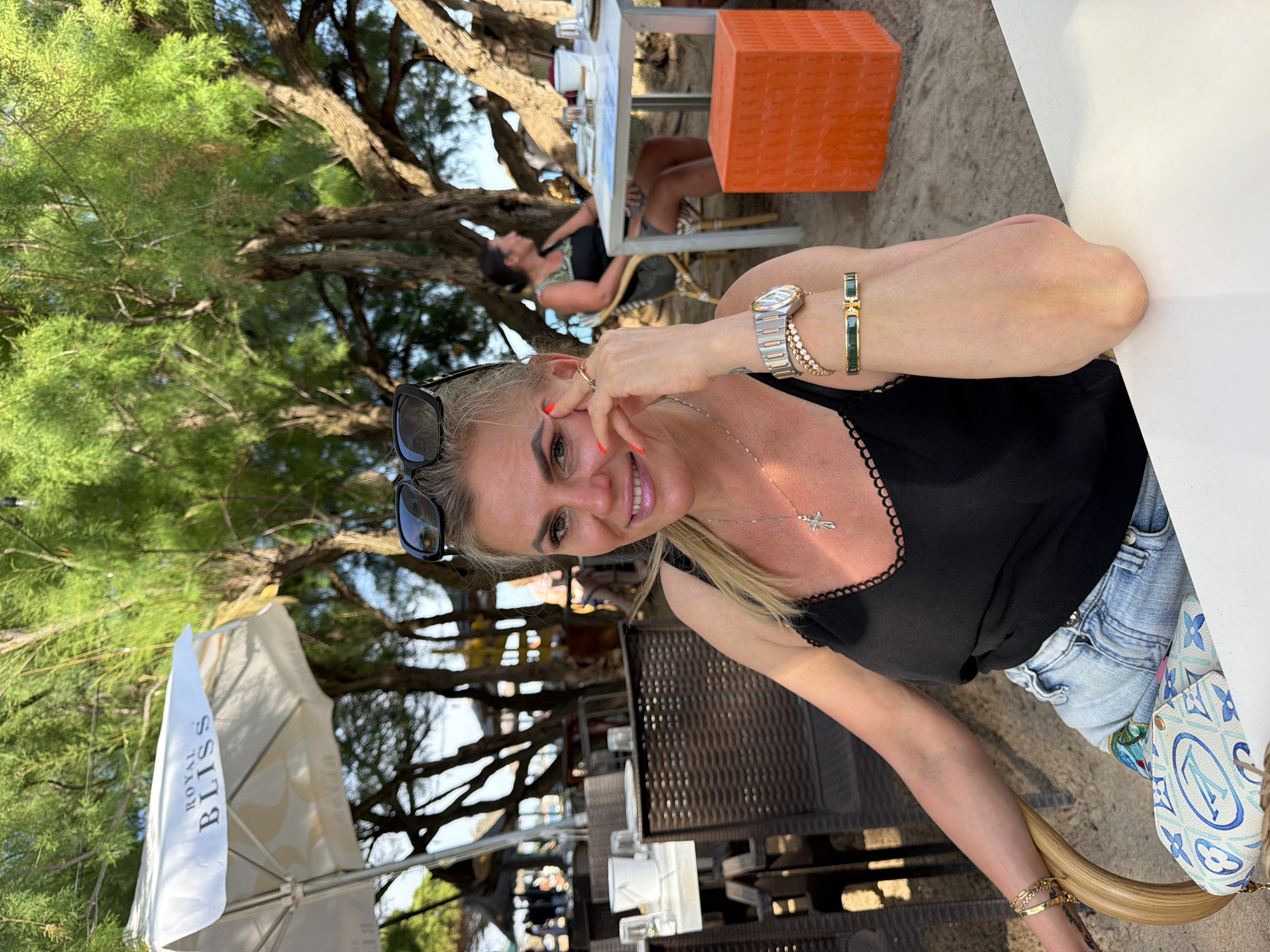 A fair-skinned woman with blonde hair and sunglasses poses at an outdoor cafe or restaurant. She is wearing a black top, denim shorts, and jewelry. She sits at a table with another table and chair in the background, where another woman is relaxing. An orange stool sits next to the table. The cafe is situated under trees, with a beach and sand in the background. A "Royal Bliss" umbrella is visible in the foreground. The location is Mont-roig del Camp, Spain.