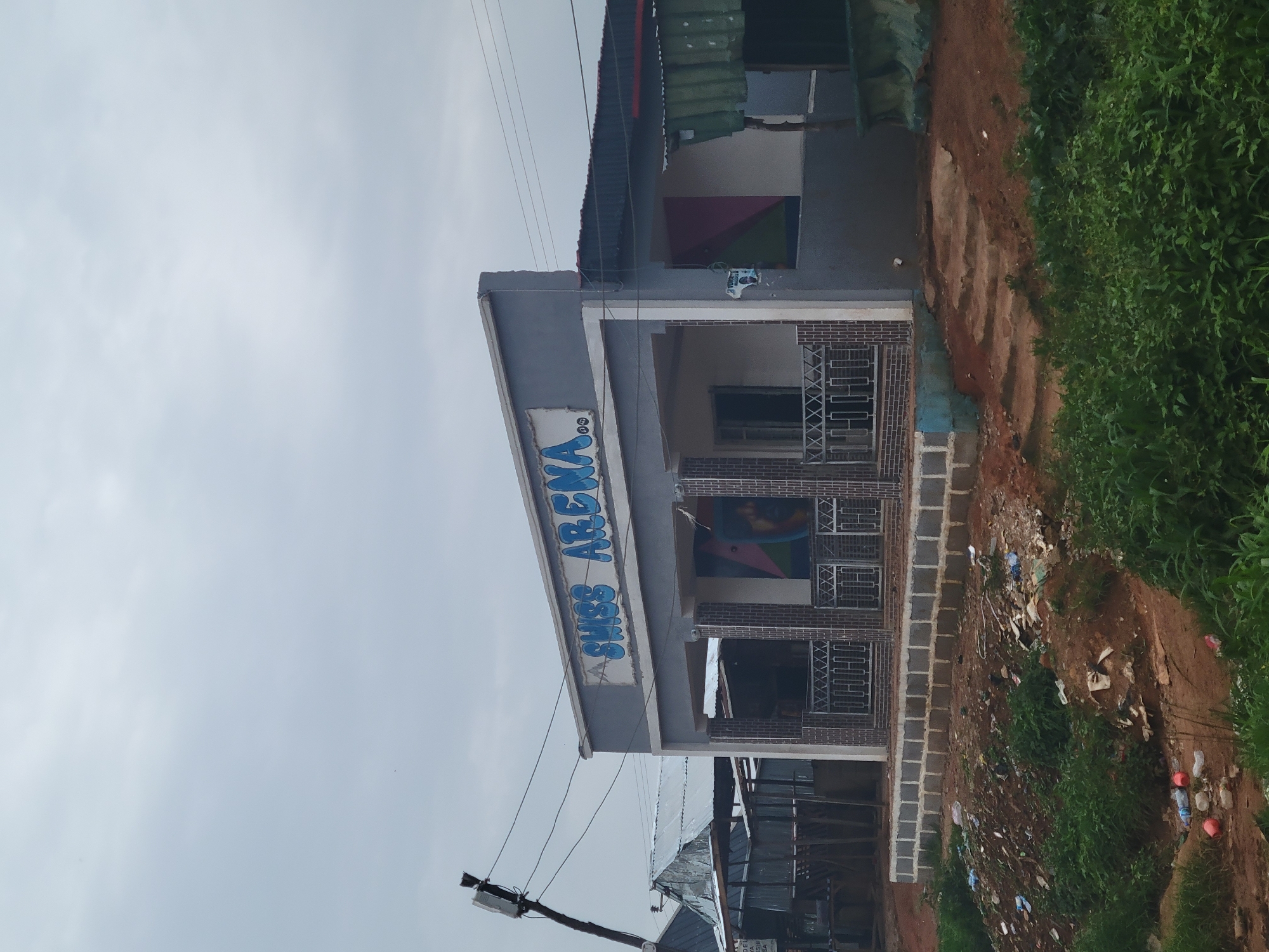 In Unknown, Unknown, a light grey building with the words "SWISS ARENA" painted in blue on its facade stands under a cloudy sky. The building has a porch with brick pillars and metal railings. To the left of the building, there are other structures with corrugated metal roofs. The foreground is a patch of ground with green vegetation and scattered litter, including plastic bottles and wrappers. A utility pole with wires is visible to the far left of the scene.
