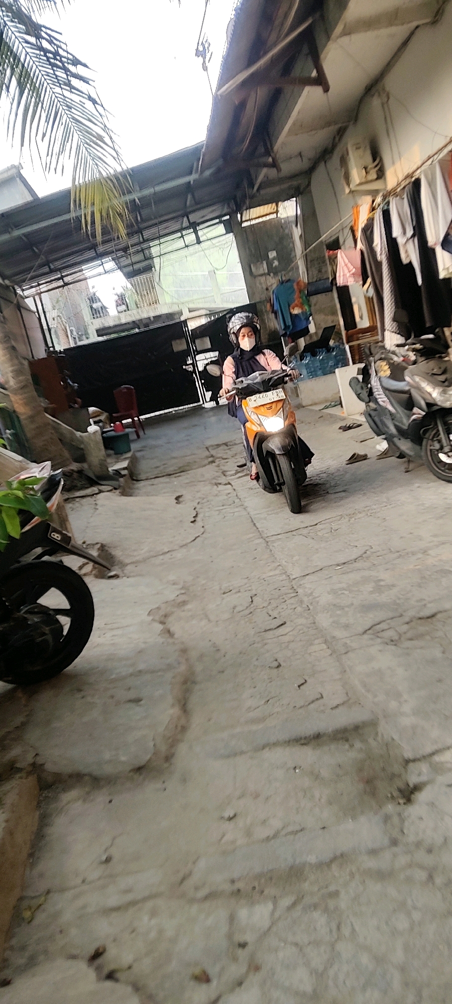 In Mekarmukti, Indonesia, a person wearing a pink shirt, dark hijab, and a white mask rides an orange and black motorcycle down a cracked concrete pathway. The motorcycle has a license plate that reads "B 3440 FGH". The pathway is lined with buildings and a covered walkway, with laundry hanging on a clothesline on the right side of the frame. There are other motorcycles parked along the path, and some greenery is visible in the foreground. The scene appears to be an outdoor alleyway or courtyard in a residential or commercial area.