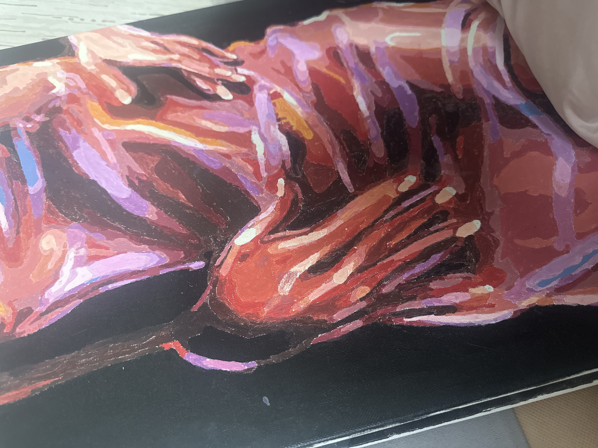 The image shows an oil painting of two figures on a black canvas. The figures are rendered in shades of red, purple, pink, and brown. The visible hands are close to each other, with a slightly visible white edge of the canvas in the bottom right corner, and a blurred white object in the upper right corner. The location of Высокий, Russia is not apparent in the image.