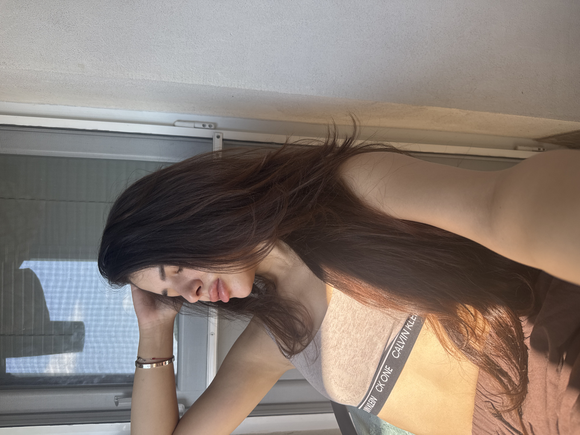 An image captures a female individual indoors, in Plovdiv, Bulgaria, positioned to the left of a white-framed window. Strong natural sunlight streams in from the left, illuminating her face, torso, and hair. The woman has long, layered dark brown hair that cascades over her left shoulder and chest. Her head is tilted back and slightly to the right, with her eyes closed and her lips full. Her right arm is bent with her hand resting near her hairline, revealing a silver-colored bracelet with visible screw details and a thin red string on her wrist. She wears a light beige Calvin Klein "CK ONE" sports bra with a black elastic band, and brown-colored pants or shorts are visible at the bottom of the frame. The window features a mesh screen, and beyond it, partially drawn blinds and a clear blue sky are visible. A textured, off-white wall occupies the right side of the frame.