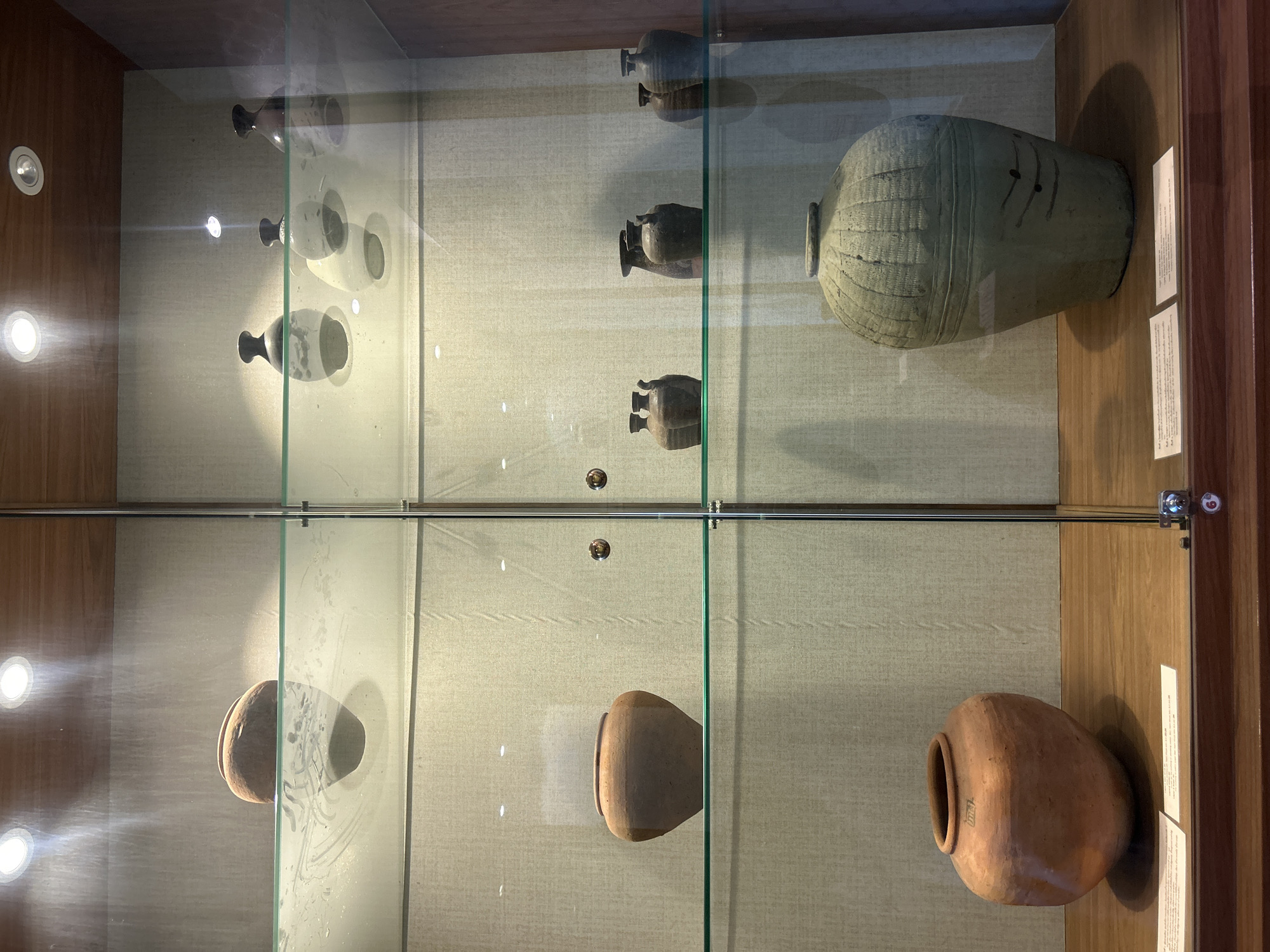 The image shows a display case containing several ancient pottery artifacts. The display case has glass shelves and is lit from above. On the top shelf, there are three small, dark vases with rounded bodies and narrow necks, and two lighter-colored, bulbous vases. On the middle shelf, there is a large, reddish-brown earthenware pot with a wide opening and a rounded base on the left. To the right of this pot, there are two small, dark vases with handles, followed by two taller, dark vases, and then two more dark vases. On the bottom shelf, on the left, there is a large, round, reddish-brown earthenware pot with a visible inscription. On the right, there is a very large, pale green, textured ceramic vessel with horizontal lines and a rounded lid. White labels are placed on the wooden surface in front of the pottery on the bottom shelf. The background of the display case is a textured, light-colored fabric. The overall setting appears to be a museum or historical exhibition. The location is Thai Ko Subdistrict, Thailand, likely within a museum context.