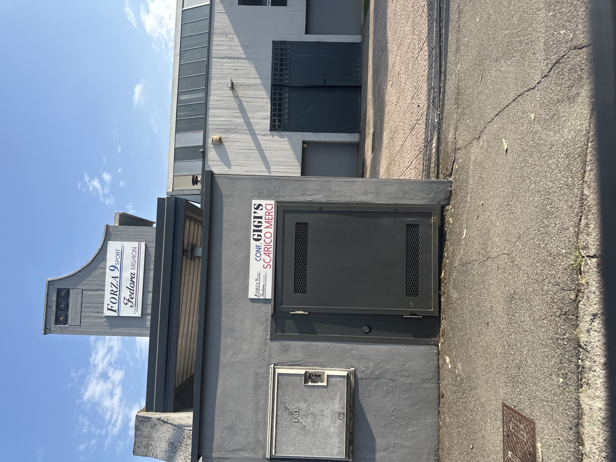 The image shows an exterior view of a commercial building with two signs. The larger sign, mounted on a grey concrete structure, reads "FORZA 9 SPORT Fedora FASHION" in a stylized font. Below this, a smaller white sign on a grey wall displays "FORZA 9 SPORT Fedora FASHION". To the right of this sign is another white sign with black text that reads "CONF. GIGI'S SCARICO MERCI". Adjacent to the signs on the left is a grey utility box with "ENEL" printed on it. A dark grey metal door with a gridded section is visible to the right of the utility box. The building's exterior is predominantly grey concrete and white panels, with a large set of dark grey double doors in the background. The ground in front of the building is paved with patterned tiles. The sky above is blue with scattered white clouds. This scene is located in Rudiano, Italy.