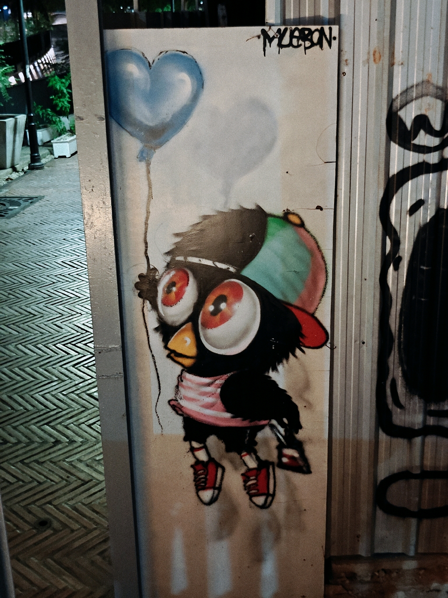 A street art mural is depicted on a light-colored panel adjacent to a corrugated metal wall. The mural features a stylized black bird with large, red-rimmed eyes, a yellow beak, and a striped pink scarf. The bird wears a cap with green, red, and blue sections and red sneakers with white soles. It is holding onto a string attached to a light blue balloon shaped like a heart. Above the balloon, black graffiti lettering reads "MLEBON". To the left of the mural, a sidewalk with a patterned texture is visible, leading towards a dimly lit area with street lights and greenery. The scene appears to be an outdoor urban environment.