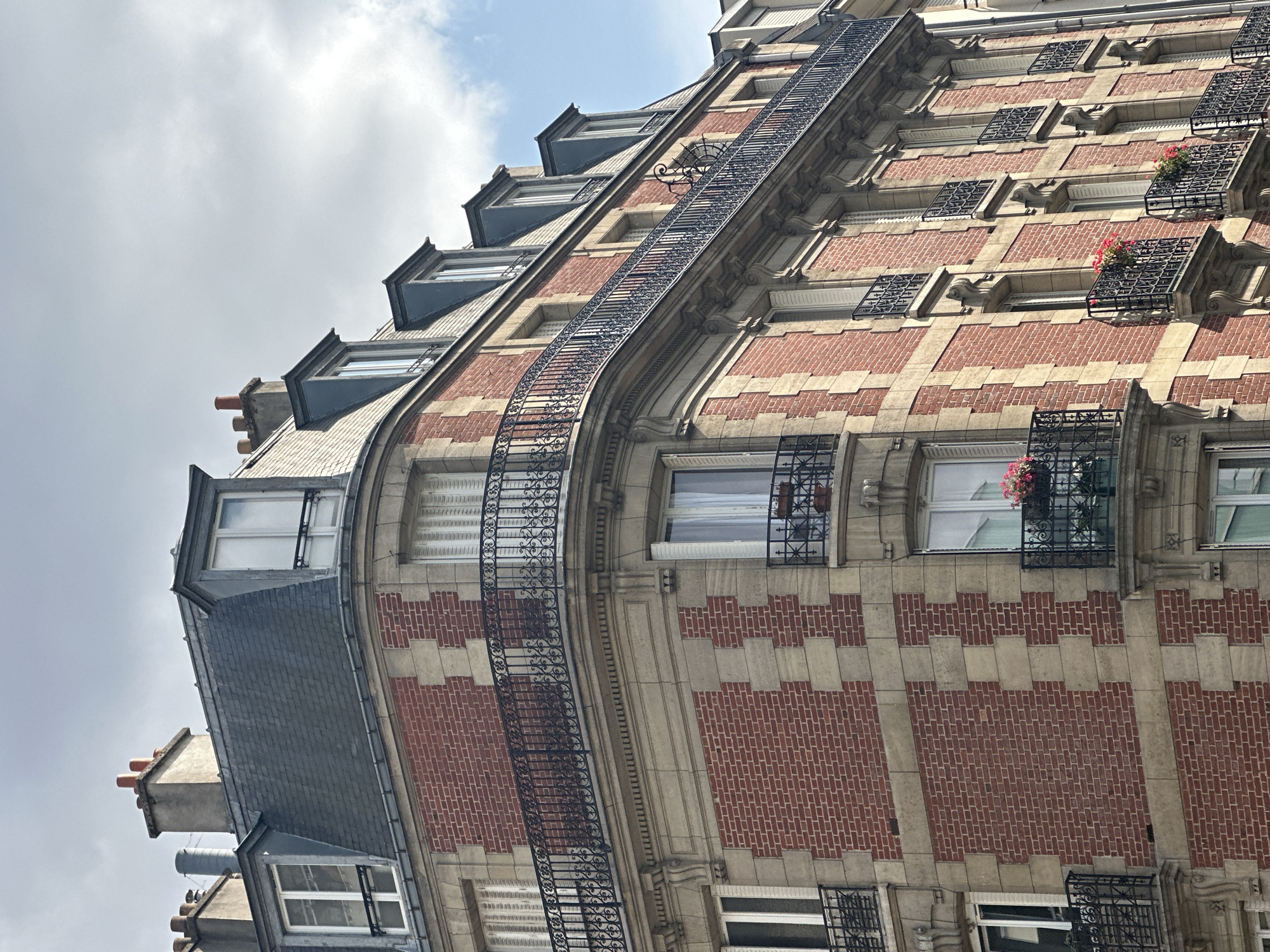 This is a close-up, low-angle outdoor shot of the exterior of a multi-story building in Paris, France. The building features a red brick facade with light-colored stone accents and decorative trim. Several windows are visible, many with white shutters and some with black wrought-iron balconies. A prominent mansard roof with dormer windows and chimneys caps the structure. The sky, visible at the top of the image, is cloudy with patches of blue. No people or animals are present in the frame.