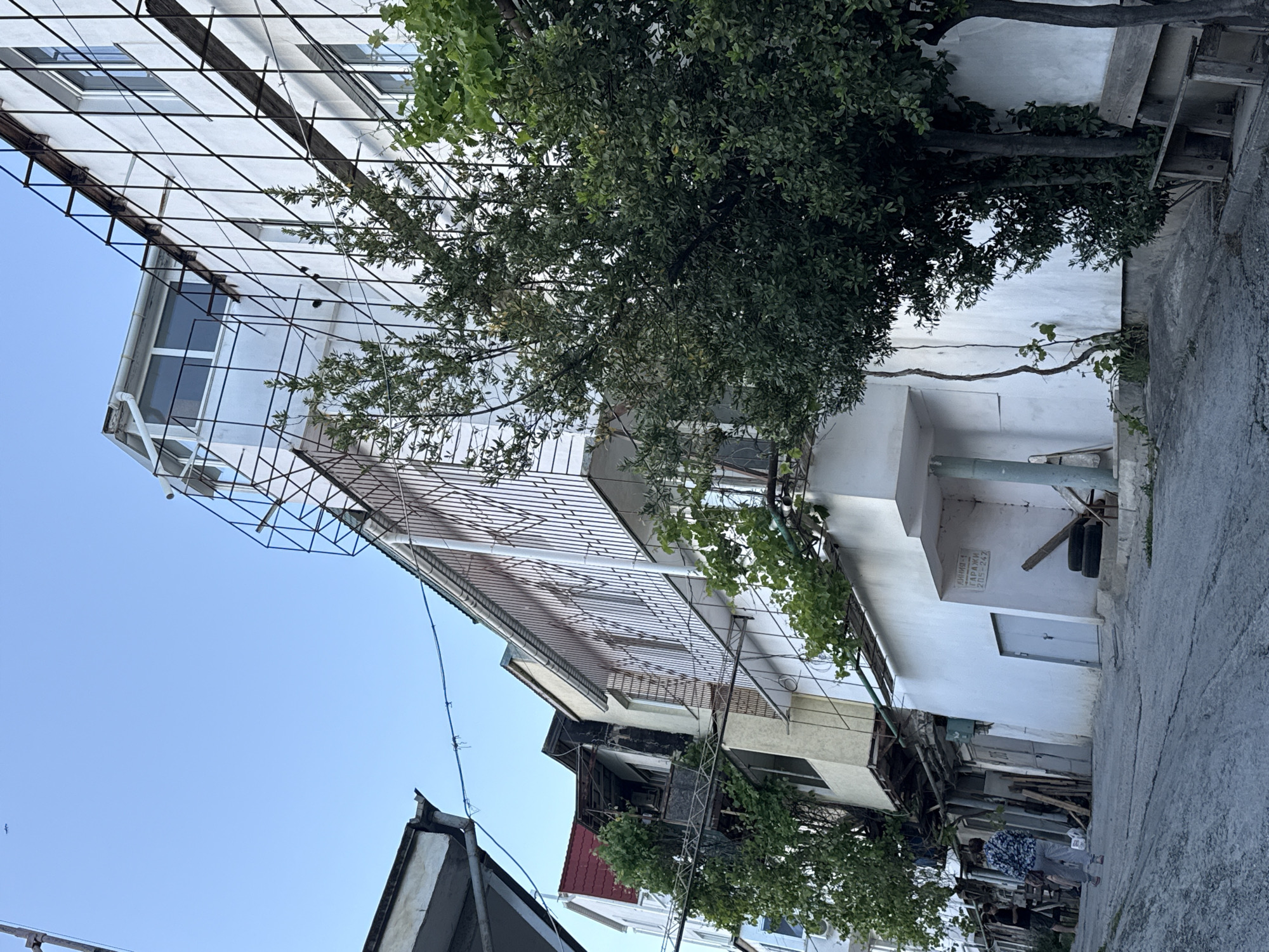 The image depicts a narrow street in an urban setting, likely in Unknown, Unknown. Buildings line the street, with white facades on some and varying architectural styles. The sky is visible above, a clear blue. Trees with dense foliage are present, partially obscuring the buildings. Electrical wires and metal structures are visible near the buildings. In the foreground, the street pavement is cracked and uneven. Further down the street, a person is visible. The scene appears to be taken during daylight hours.