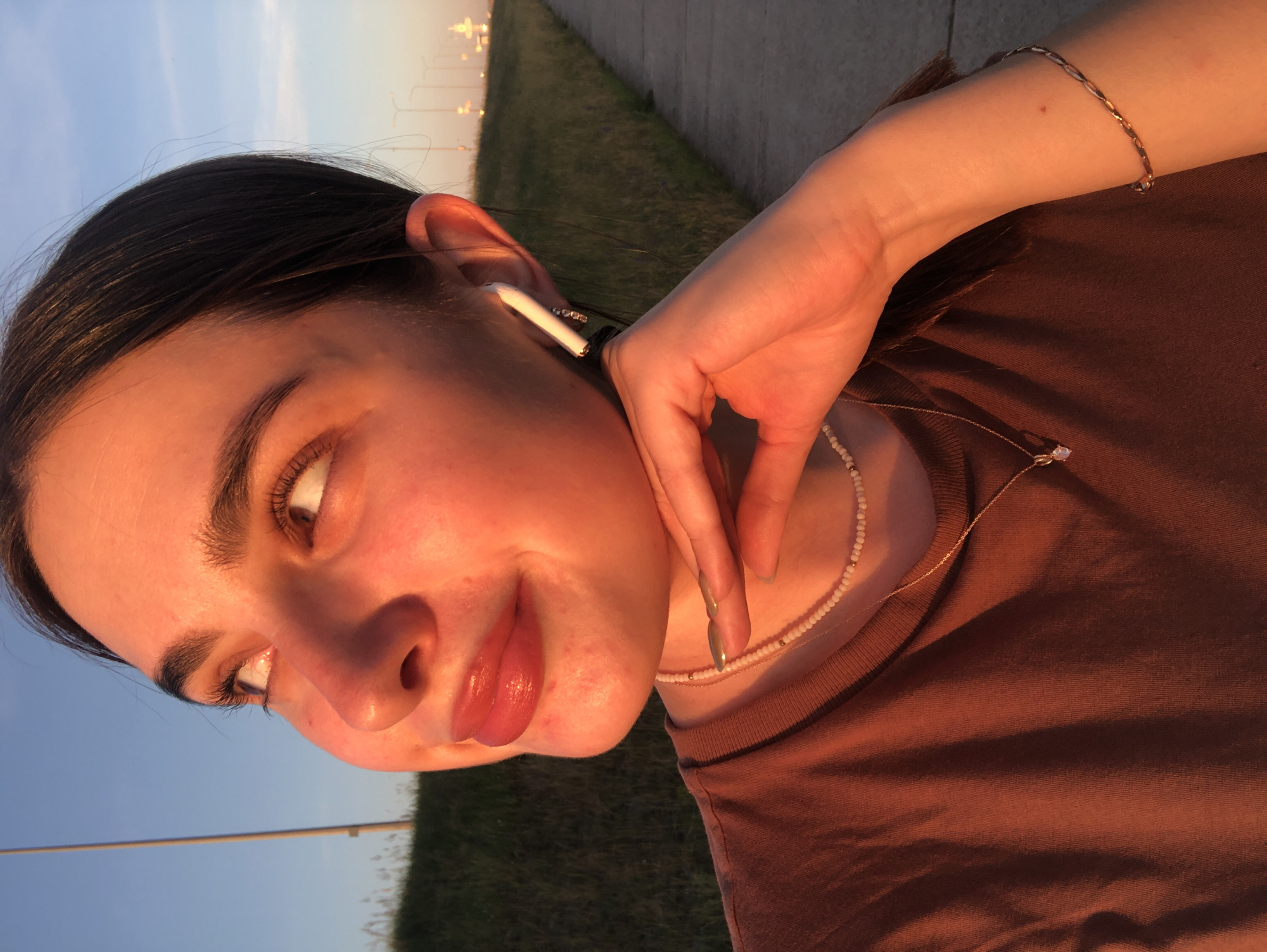 The image shows a close-up of a young woman outdoors during what appears to be sunset. The golden light casts a warm glow on her face, highlighting her features: her eyes are looking upwards, her lips are slightly parted, and her chin rests on her hand. She is wearing a brown t-shirt, a beaded necklace, and a delicate gold necklace with a pendant. An AirPod is visible in her right ear. The background suggests a suburban or semi-urban environment with a grassy embankment, a paved walkway, and a row of streetlights in the distance. The sky is a clear blue, transitioning into hues of orange and pink near the horizon. The setting evokes a sense of peace and contemplation. The image was taken in Noviy, Russia.