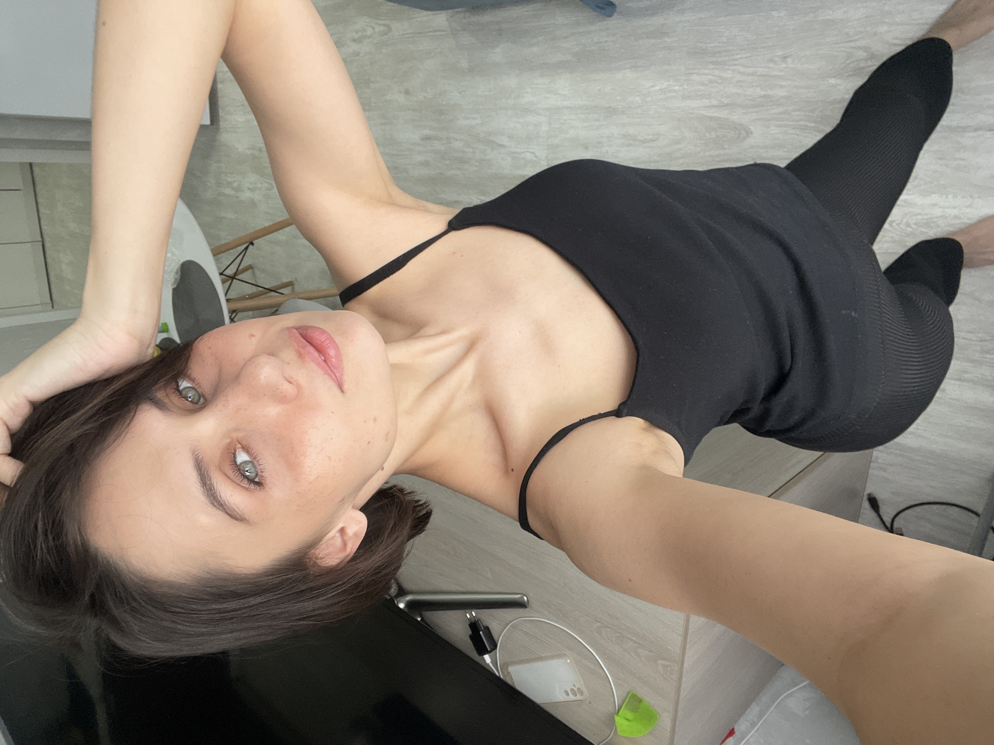 A young woman is depicted lying on her back, looking directly at the camera with a calm, neutral expression and a slight smile. She has dark hair, prominent green eyes, and visible freckles across her face. She is wearing a simple black spaghetti-strap tank top and black ribbed leggings. One arm is raised and positioned behind her head, indicating a relaxed pose, and her bare feet are visible in the background.
The photograph is taken from a low angle, giving an upward perspective, and the entire scene appears rotated. The setting is indoors, characteristic of a home or apartment in Izhevsk, Russia. The flooring is light-colored, resembling wood or laminate. A light-toned table or desk is in the foreground, holding several items: a dark electronic screen (possibly a television or monitor), a white charging cable, a black wall charger, a white phone case (suggesting a Samsung model), and a small green object. In the background, hints of other household items, including parts of a chair or scale, are visible. The lighting is natural, suggesting the image was captured during the daytime. No text is visible in the image.