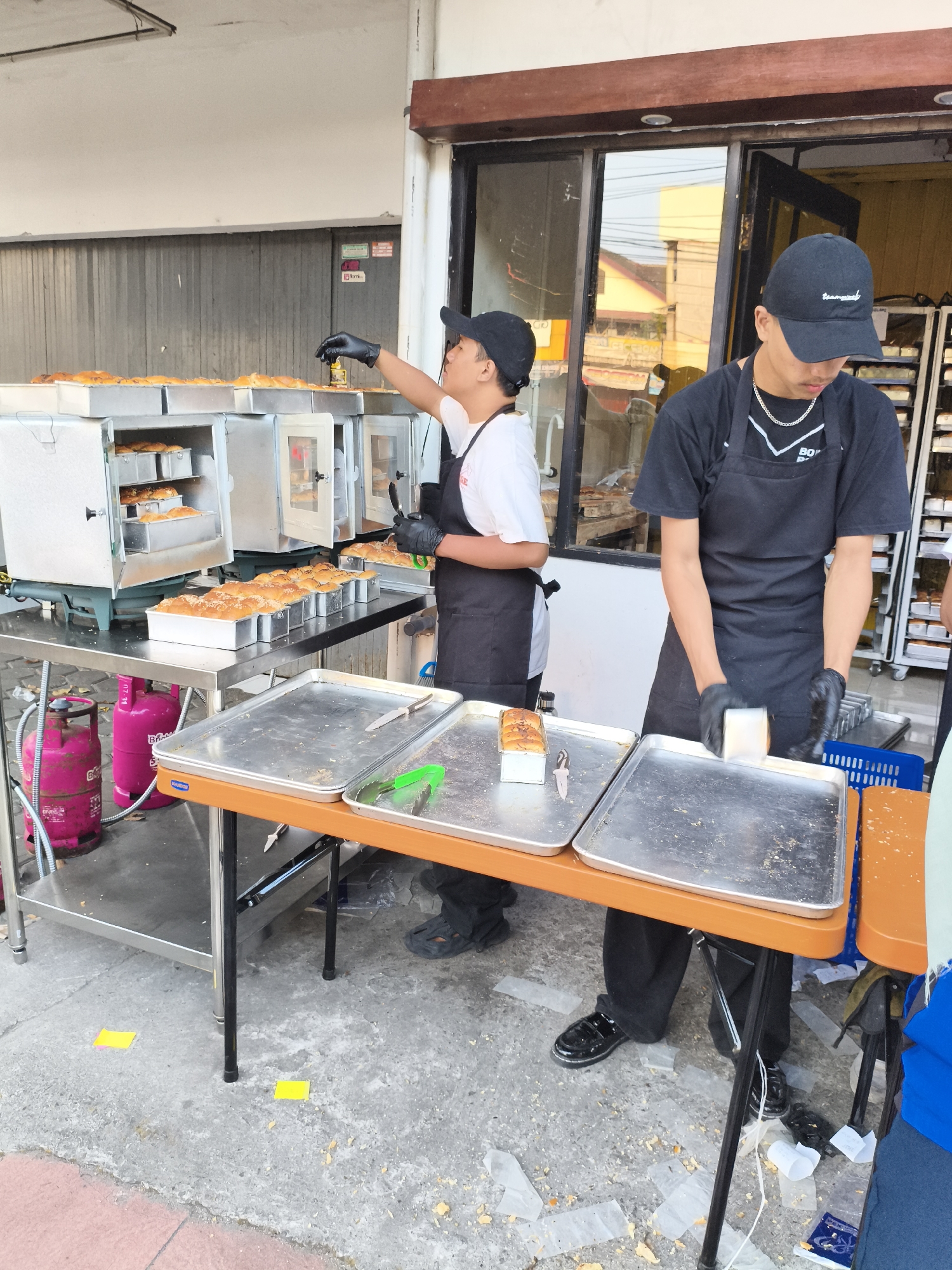 Two men, wearing black aprons and black baseball caps, are in the process of preparing bread at an outdoor stall. One man, on the left, is wearing a white shirt and black apron, and is reaching into an open oven with gloved hands, seemingly to retrieve baked bread. The other man, on the right, is wearing a black shirt and black apron, and is using gloved hands to arrange bread on a metal tray. Multiple ovens are visible behind them, filled with bread in baking pans. Several metal trays with baked bread are also displayed on tables in front of the stall. Two pink gas cylinders are located to the left of the stall. The ground is paved with concrete and shows some scattered debris. Yellow sticky notes are visible on the ground in front of the stall. The background shows the facade of a building with windows and a doorway. The setting appears to be a street in the city of Magelang, Indonesia, based on the style of the building and the presence of the gas cylinders, commonly used for street vendors in Indonesia.