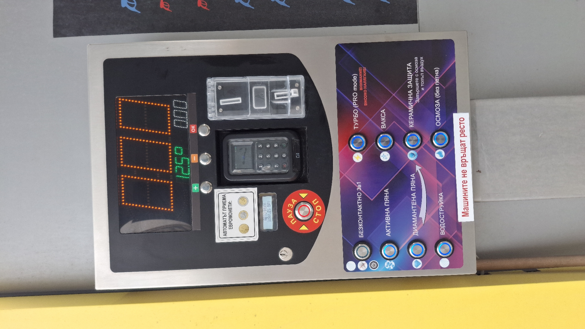 The image shows the control panel of a self-service car wash station. The panel is made of stainless steel and black plastic. It includes a digital display showing "12.50" in green, with "0.00" displayed above in orange. There are buttons for various washing options in Bulgarian: TURBO (PRO mode), VAKSA (Wax), KERAMICHNA ZASHTITA (Ceramic Protection), OSMOZA (Osmosis), BEZKONTAKTNO 3v1 (Contactless 3 in 1), AKTIVNA PYANA (Active Foam), DIAMANTENA PYANA (Diamond Foam), and VODOSTRUYKA (Water Jet). There's a coin slot with the inscription "AVTOMAT"T PRIEMA EVROMONETI" (The machine accepts Euro coins), a keypad, and a red "PAUZA/STOP" (Pause/Stop) button. A label reads "Машините не връщат ресто", translated as "The machines do not return change." The background shows a blurred image. Given the Bulgarian language on the panel, it is likely located in Bulgaria. Without further information, the exact location in Varna cannot be determined.