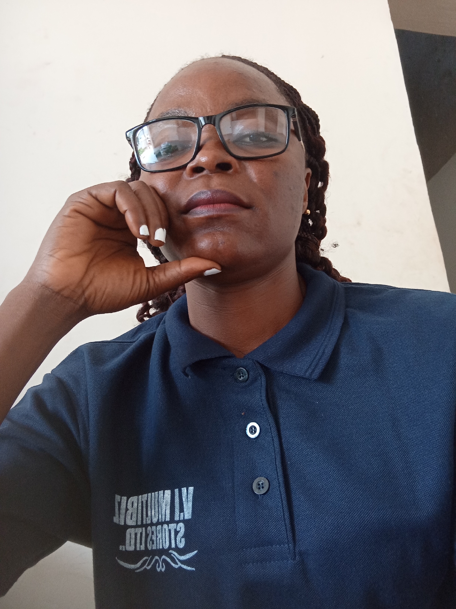 A woman wearing glasses and a dark blue polo shirt is looking directly at the camera. She has her hand resting on her chin, with her fingers splayed. Her nails are painted white. She has dark hair braided into long strands that fall over her shoulders. The background is a plain, light-colored wall. The text on her shirt appears to be in white lettering and is partially obscured by the camera angle. The image seems to have been taken indoors, with neutral lighting. The woman's expression is neutral. The scene suggests a casual, everyday moment. The text on the shirt is partially legible and appears to be reversed, possibly due to the camera's perspective. It reads "STORES LTD" with some other characters that are difficult to discern. There are also decorative lines beneath the text.