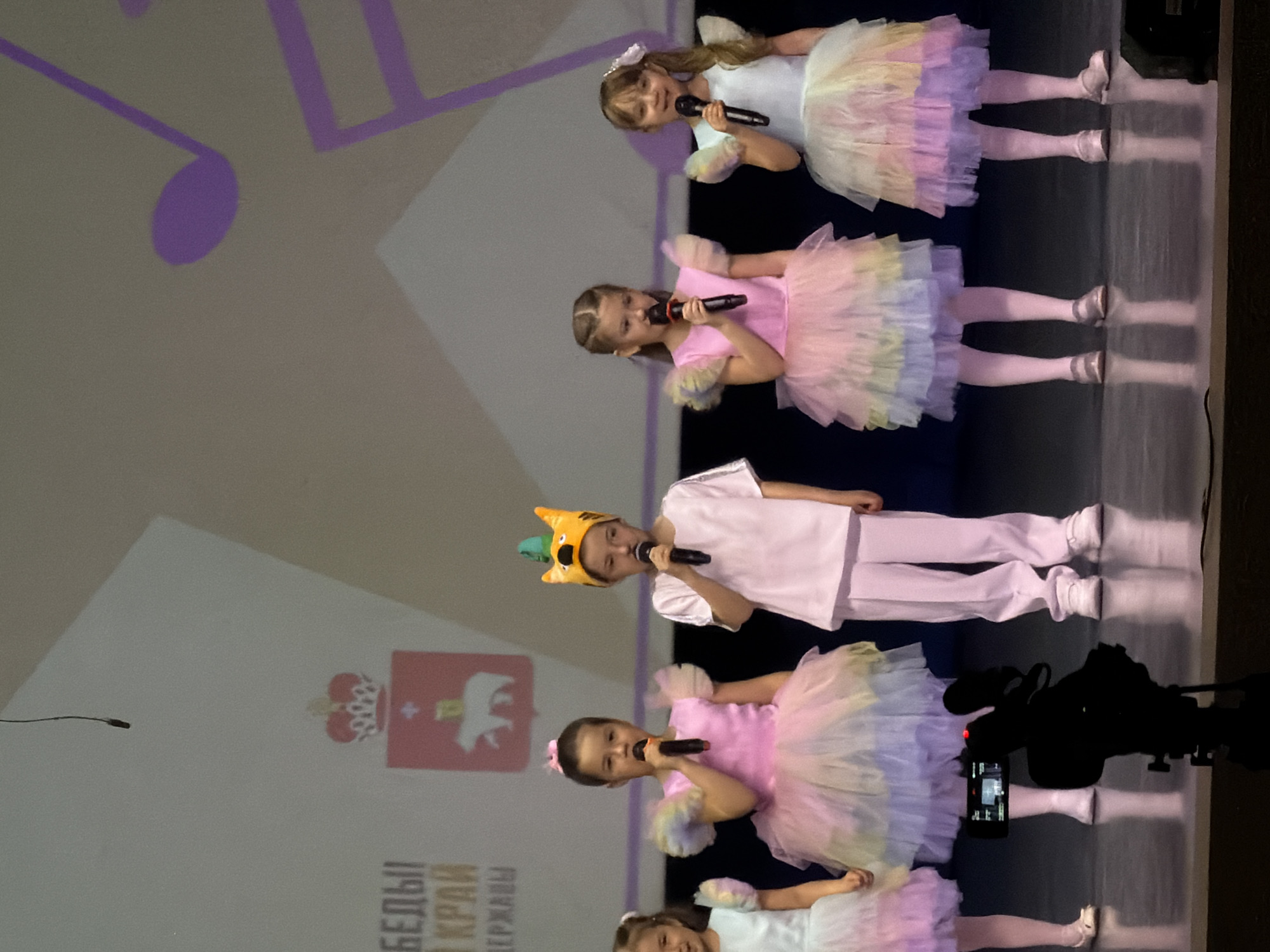 The image shows five young girls performing on a stage. Four of the girls are wearing colorful tutus and holding microphones. The middle girl is wearing a white outfit and a cartoon hat, also holding a microphone. They are standing in a line and appear to be singing. Behind them is a screen displaying musical notes and the coat of arms for the city of Perm, Russia, suggesting the performance is taking place in Perm. A camera setup is visible on the right side of the frame.