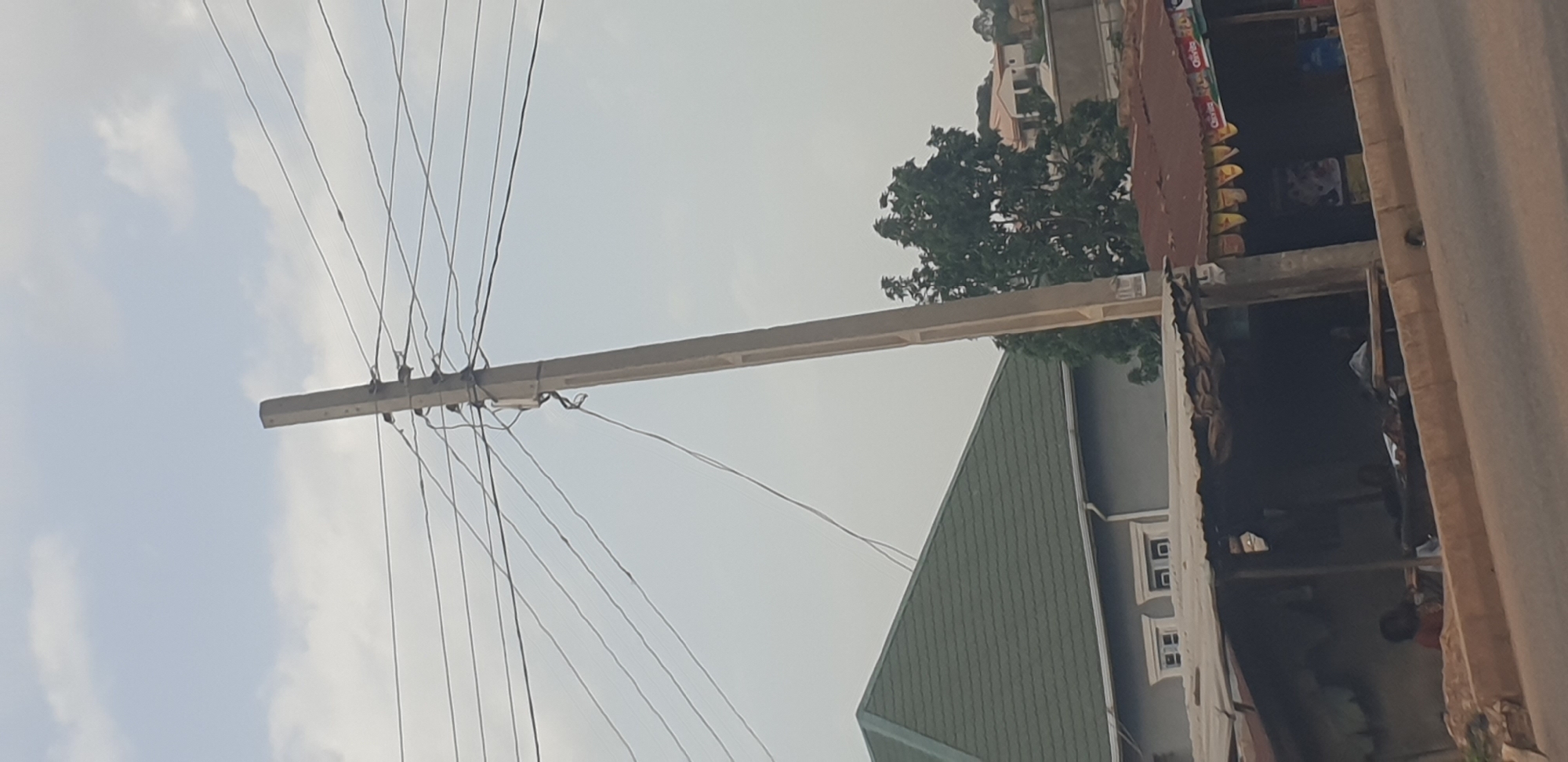 The image captures a view along a street in Barakin Lerawi, Nigeria, dominated by a tall, concrete utility pole laden with multiple electrical wires. The pole is positioned slightly to the right of the frame and is the central focus of the upper portion of the image, with wires extending horizontally across the sky. Below the utility pole, a weathered, corrugated metal roof covers a roadside structure, appearing to be a small shop or stall. This structure is built against a sloping embankment. To the left of this stall, a house with a prominent, dark green, gabled roof is visible. The house has light grey walls and features multiple windows with white framing. Further in the background, dense green trees partially obscure other buildings and structures, suggesting a suburban or semi-urban environment. The sky above is a pale, hazy blue with scattered, wispy clouds. The foreground shows a paved road and a concrete curb. There are a few indistinct figures visible within the shaded area of the roadside stall, suggesting human presence but their activities are not clearly discernible.