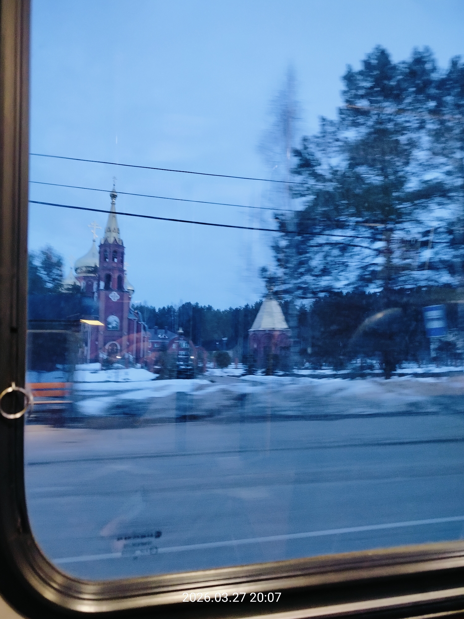 The image provides a view through a vehicle window at 8:07 PM on March 27, 2026, depicting an evening scene in Chaikovsky, Perm Krai, Russia. The primary subject is the Church of St. George (Храм Георгия Победоносца), a large, red-brick Orthodox church featuring multiple golden onion domes and crosses, positioned in the middle ground to the left. To the right of the main church, a smaller brick structure with a light-colored conical roof is visible. The ground is partially covered in patches of snow, indicative of a late winter or early spring environment. A dark asphalt road, appearing somewhat reflective or wet, occupies the lower portion of the frame. A dense, blurred treeline, consisting of both evergreen and bare deciduous trees, forms the background. Several dark power lines stretch horizontally across the upper middle section of the image. The immediate foreground shows the dark frame of the vehicle's window and a metal ring on the left edge. The entire outdoor scene is characterized by significant motion blur, consistent with a photograph taken from a moving vehicle. No individuals or specific actions are discernible within the frame.