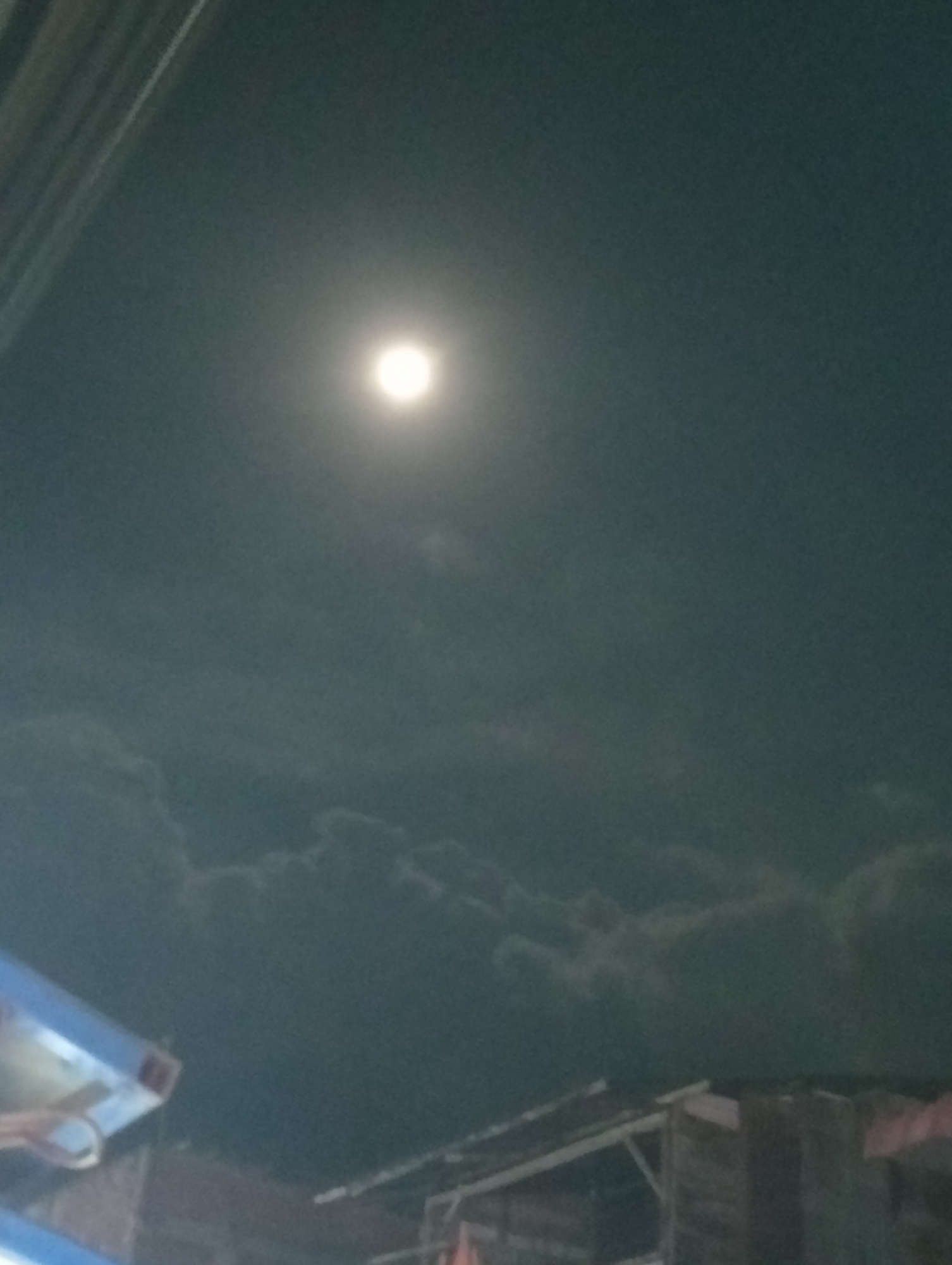 At night in Pulorayeuk, Indonesia, a bright moon shines above a low-hanging layer of clouds. The view is from below, looking up. A roof structure is visible in the lower part of the frame. The overall lighting is dim due to the night setting.