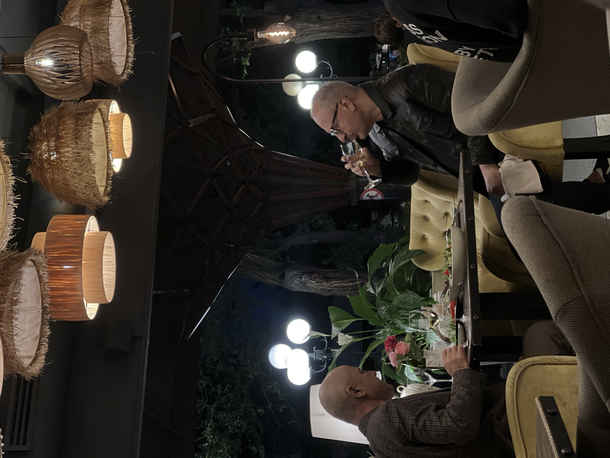 The image depicts an outdoor dining scene in Yerevan, Armenia, likely at a restaurant or café. Two men are seated at a table, engaged in conversation. One man, wearing glasses and a dark jacket, is drinking from a wine glass. The other man, bald and wearing a patterned jacket, is also sitting at the table. The table is set for a meal with glassware, plates, and floral decorations. The surroundings include outdoor lighting fixtures, such as pendant lights and street lamps with multiple globes. There is lush greenery in the background, suggesting an outdoor patio or garden setting. Seating consists of cushioned chairs.