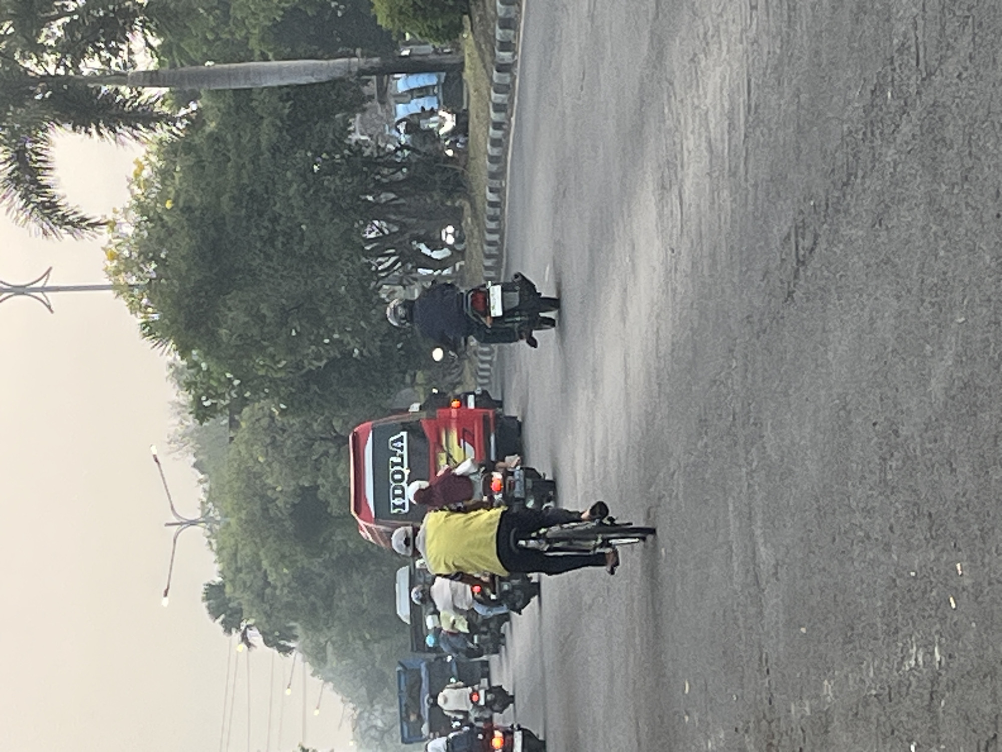 The image depicts a street scene in Kludan, Indonesia, likely during daylight hours with overcast conditions. A red bus with "IDOLA" written on its rear is stationary in the middle lane, surrounded by other vehicles. Several individuals are riding motorcycles, and one person is on a bicycle, all moving in the same direction along the asphalt road. The road is bordered by a sidewalk and trees, with power lines visible in the background. The overall atmosphere is one of moderate traffic and urban activity.