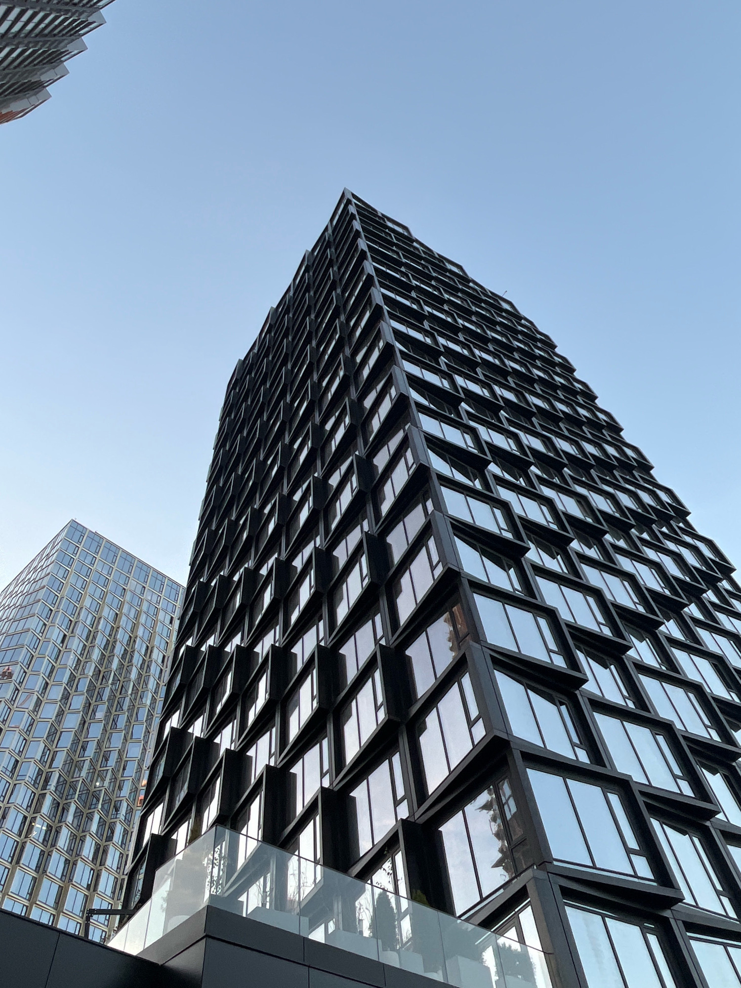 This image shows a low-angle view of two modern skyscrapers against a clear blue sky. The skyscraper in the foreground is tall and slender, with a dark facade featuring a grid of windows that are set at an angle, creating a textured, diamond-like pattern. The windows reflect the sky and appear to be tinted. A glass balcony or terrace is visible on a lower level of this building, with faint reflections of trees and building structures.
To the left and slightly behind the foreground building is another skyscraper with a lighter, more uniform facade. This building also has a grid pattern of windows, but these are arranged more conventionally and reflect the sky with a more even, shimmering effect. The overall impression is one of contemporary urban architecture, with clean lines and reflective surfaces. The scene is devoid of any people or vehicles, focusing solely on the architectural elements of the city. The sky is bright and clear, suggesting daytime.
The buildings depicted are the **Neva Towers** in Moscow, Russia. The foreground building is the Neva Tower 2, and the building to the left is Neva Tower 1.