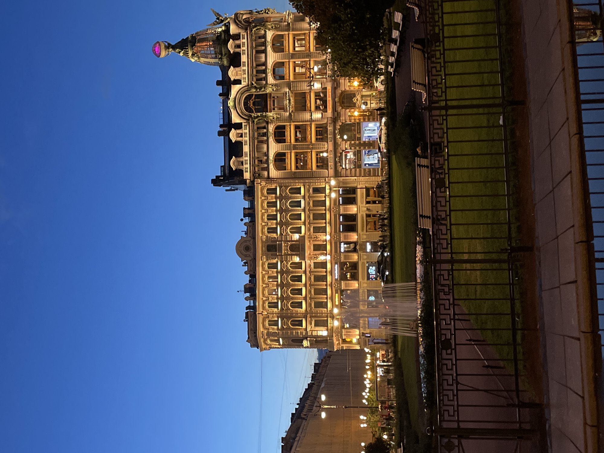 The image depicts the Singer House, also known as Dom Knigi, in Saint Petersburg, Russia, at dusk. The iconic building, illuminated by warm yellow lights, stands prominently on the left side of the frame. Its distinctive glass dome, topped with a globe adorned with a pink light, is clearly visible against the deep blue twilight sky. To the left of Singer House, another ornate building of similar architectural style is partially visible.
In the foreground, a black wrought-iron fence encloses a grassy park area. Several white park benches are arranged in rows within the park. A fountain is actively spraying water in the middle ground, with its water jets creating a misty effect. Behind the fountain, a few cars are parked on the street. Pedestrians are visible walking on the sidewalk and near the building entrances. The scene conveys a sense of urban tranquility as day transitions into night.