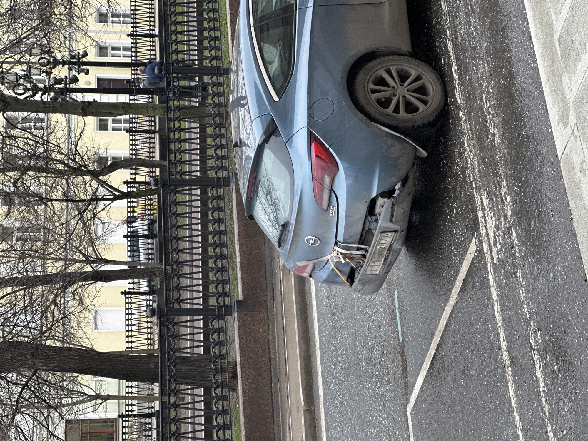 The image depicts a blue Opel Astra with significant rear-end damage parked on a wet street next to a wrought-iron fence. The license plate, partially obscured, reads "T116MX777". The vehicle's bumper is detached and secured with rope or cable. The backdrop includes bare trees and a yellow building with multiple windows. In the background, a person is sitting on a bench. The scene appears to be in Moscow, Russia.