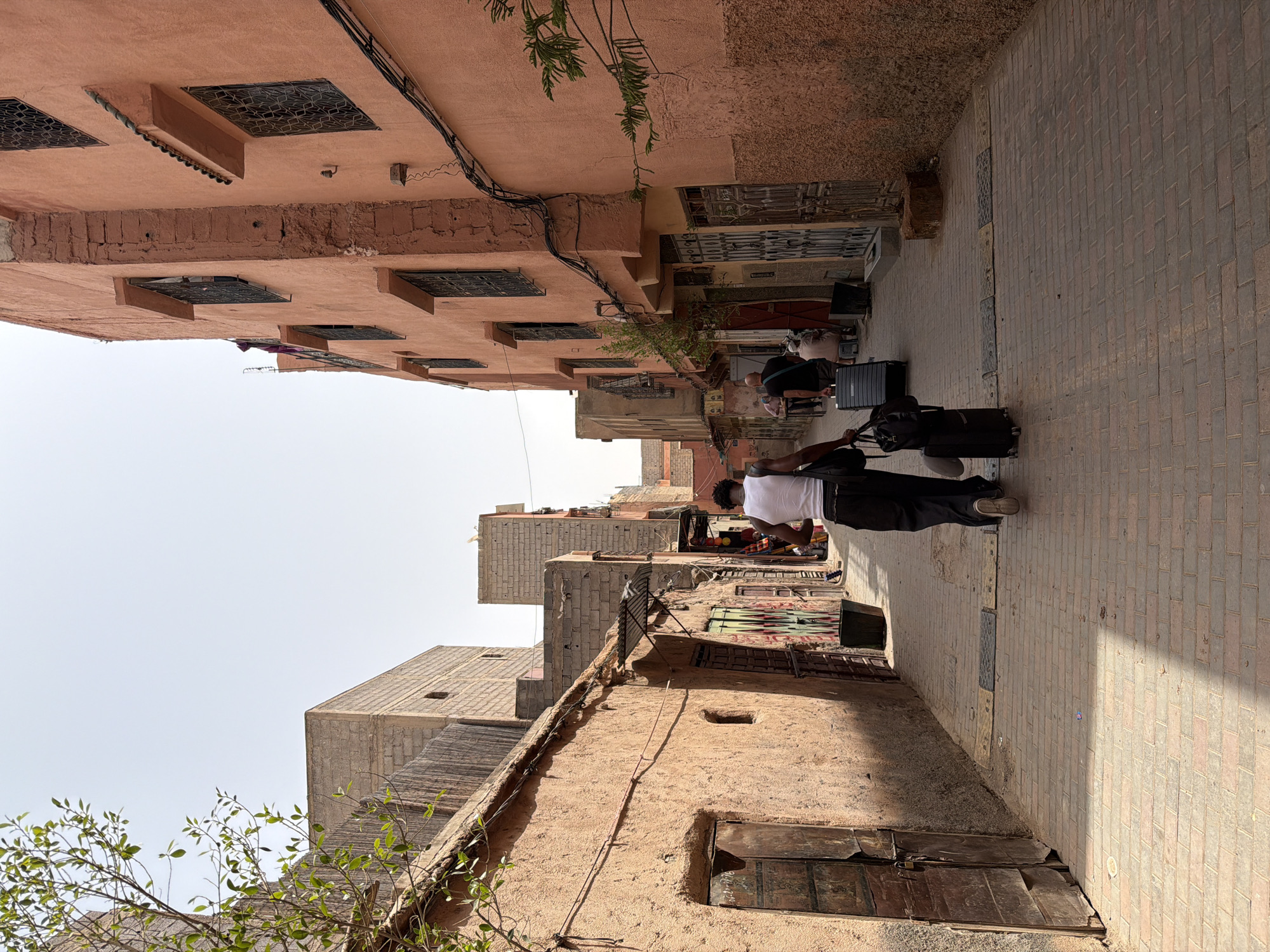 In Marrakesh, Morocco, a narrow alleyway is depicted with buildings on both sides. The left side features a textured, light brown building with a weathered wooden door and a small, arched window. The right side is dominated by an orange-hued building with multiple windows featuring dark, ornate grilles and awnings. Wires run along the exterior of this building. The ground is paved with red brick.
In the foreground, a person wearing a white tank top and dark pants is walking away from the camera, pulling a large black suitcase with wheels and carrying a black duffel bag. Further down the alley, two other individuals are seated, one on a stool and another on a bench, near a shop entrance. The overall atmosphere is that of a bustling, sunlit urban environment in Morocco.