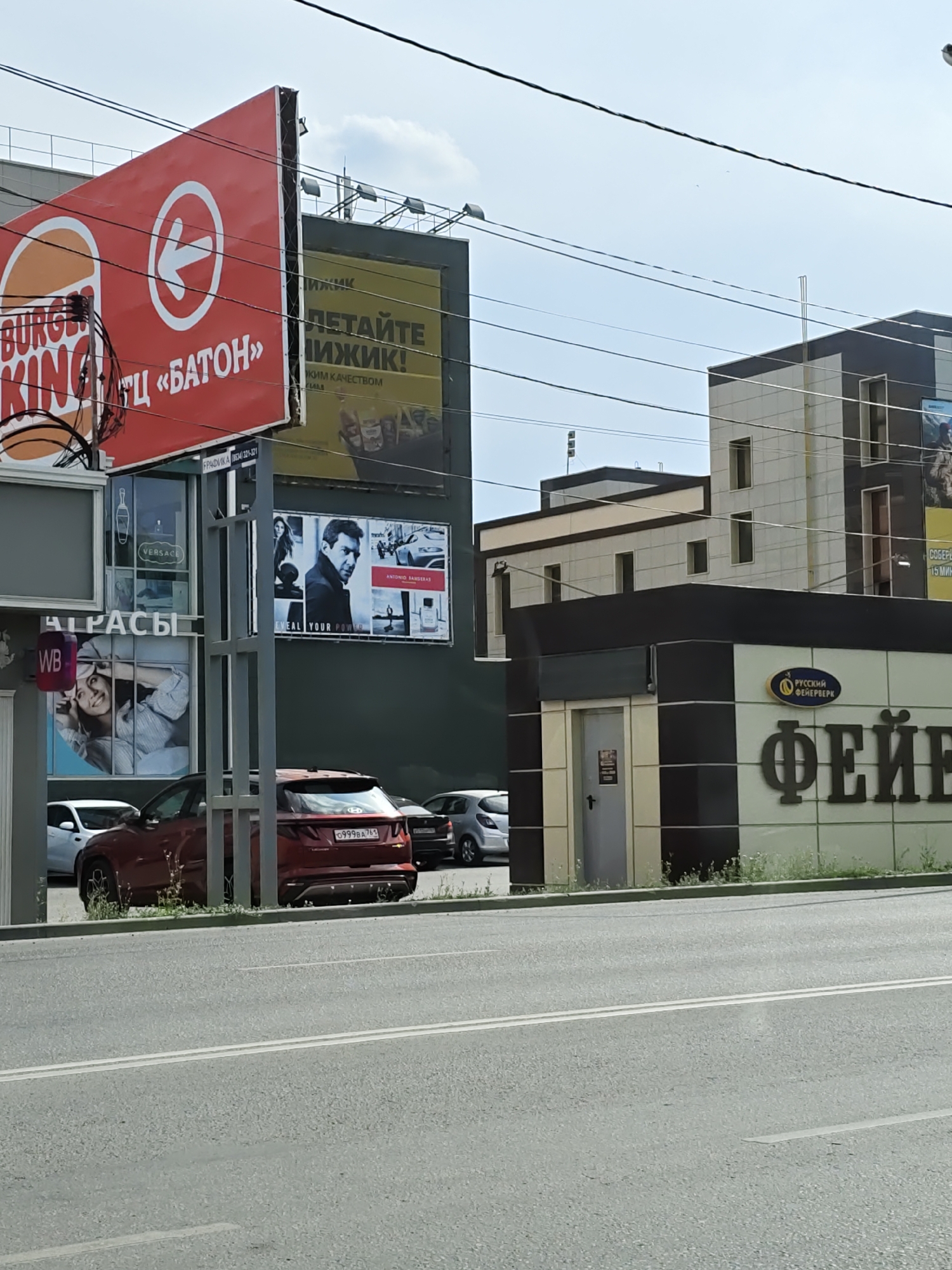 A street scene in Novocherkassk, Russia, on a clear day shows several commercial buildings and billboards. On the left, a large red Burger King billboard is visible, with the brand logo and text reading "ОТЦ «БАТОН»" (OTTS "BATON"). Below this, a section of a building displays advertisements, including one for "VERSACE" and another with a woman's face and the text "WB". To the right, a yellow billboard with Russian text is partially visible. Further right, a multi-story building with a dark and light facade stands, adjacent to a smaller structure with a dark and light tiled exterior. This smaller structure features a sign that reads "РУССКИЙ ФЕЙЕРВЕРК" (RUSSIAN FIREWORKS) in a blue oval, and large dark lettering spelling "ФЕЙЕ" (FEIE). Several cars are parked on the side of the road in front of these buildings.