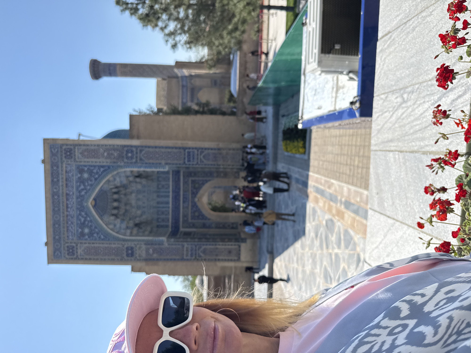 A person in a pink hat and white-framed sunglasses is taking a selfie in front of the Gur-e-Amir Mausoleum in Samarkand City, Uzbekistan. The entrance to the mausoleum is behind the person and to the left of the frame. A minaret is visible behind the mausoleum, to the left of the frame. People are visible standing and walking in front of the mausoleum's entrance. The background features a blue sky. Red flowers are present on the right side of the frame.