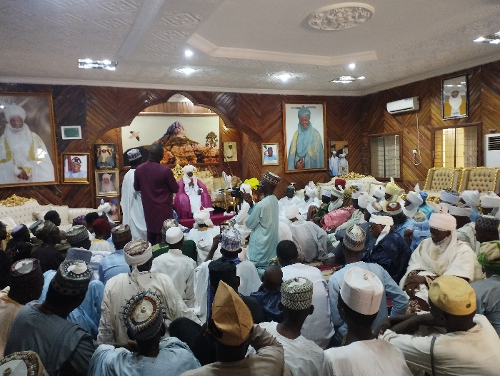 The image shows a large gathering of men in a room with wood-paneled walls and a white ceiling. The men are seated on the floor, listening to a man in a white robe and turban who is speaking from a small stage in the center of the room. There are several portraits hanging on the walls, including a large portrait of a man in traditional clothing. The room appears to be located in the Emir's Palace in Kaltungo, Nigeria, as indicated by the coordinates (latitude: 9.816, longitude: 11.315).