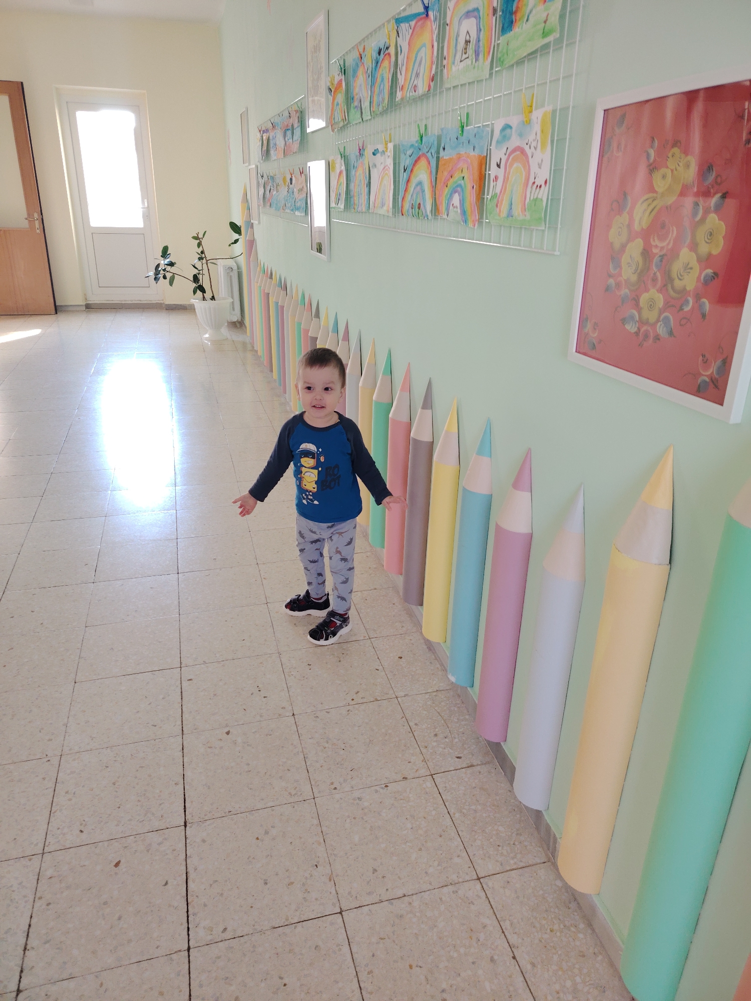 A young boy wearing a blue long-sleeved shirt with a graphic design, grey patterned pants, and black sneakers stands in the middle of a brightly lit hallway. The hallway has light-colored tile flooring and pale green walls decorated with children's artwork, including drawings of rainbows and a framed painting with floral motifs. The walls are also adorned with large, colorful, 3D pencil-shaped decorations. A white door and a potted plant are visible in the background. The photo was taken in Markovskii, Russia.