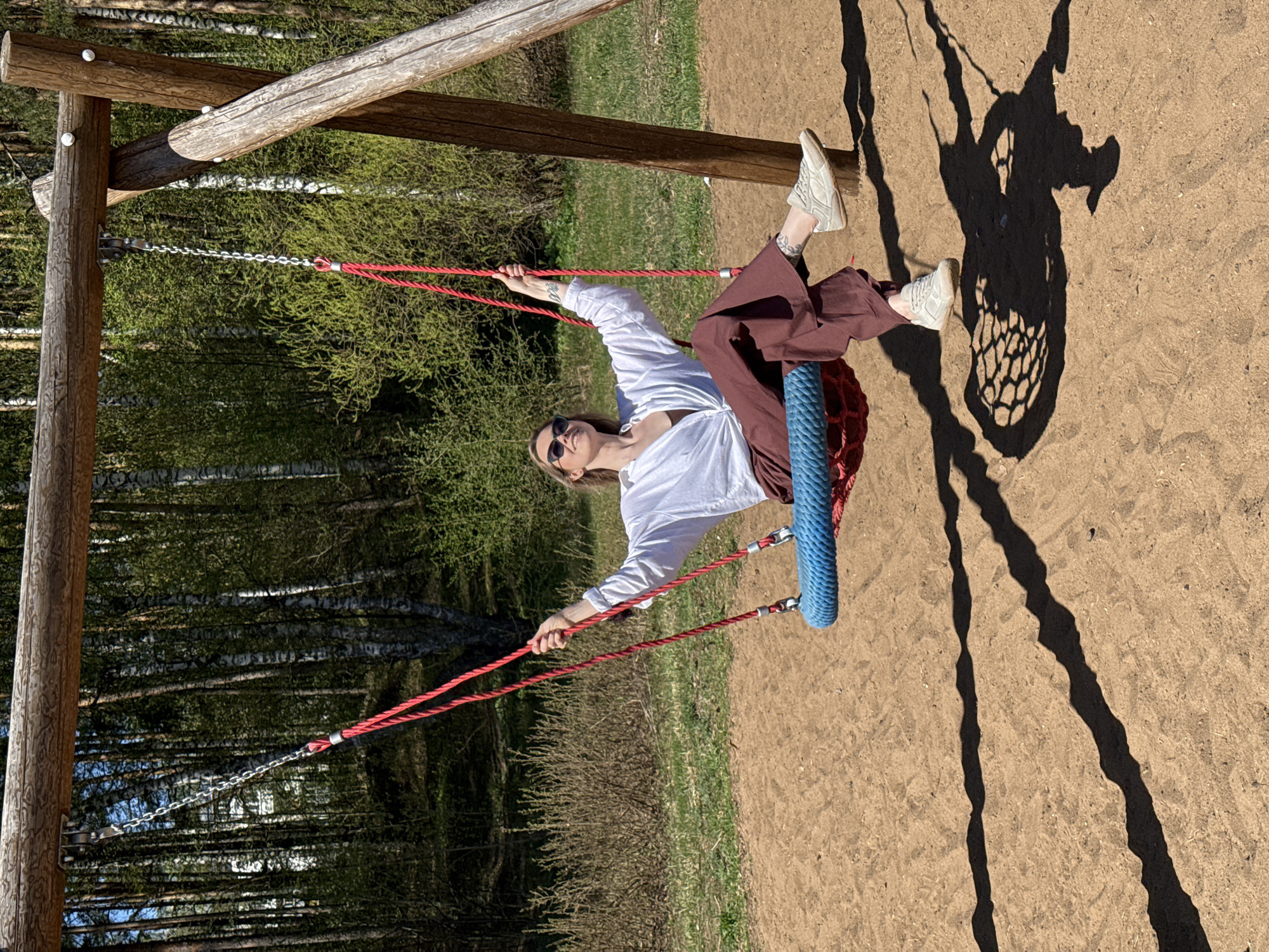 A woman sits on a tire swing in an outdoor area with trees in the background, likely located in or near the city of Chaikovsky, Russia. She wears sunglasses, a white blouse, and brown pants, and light-colored sneakers. The tire swing hangs from a wooden frame, with red ropes. The ground is sandy. A shadow of the swing and the woman is cast on the ground.