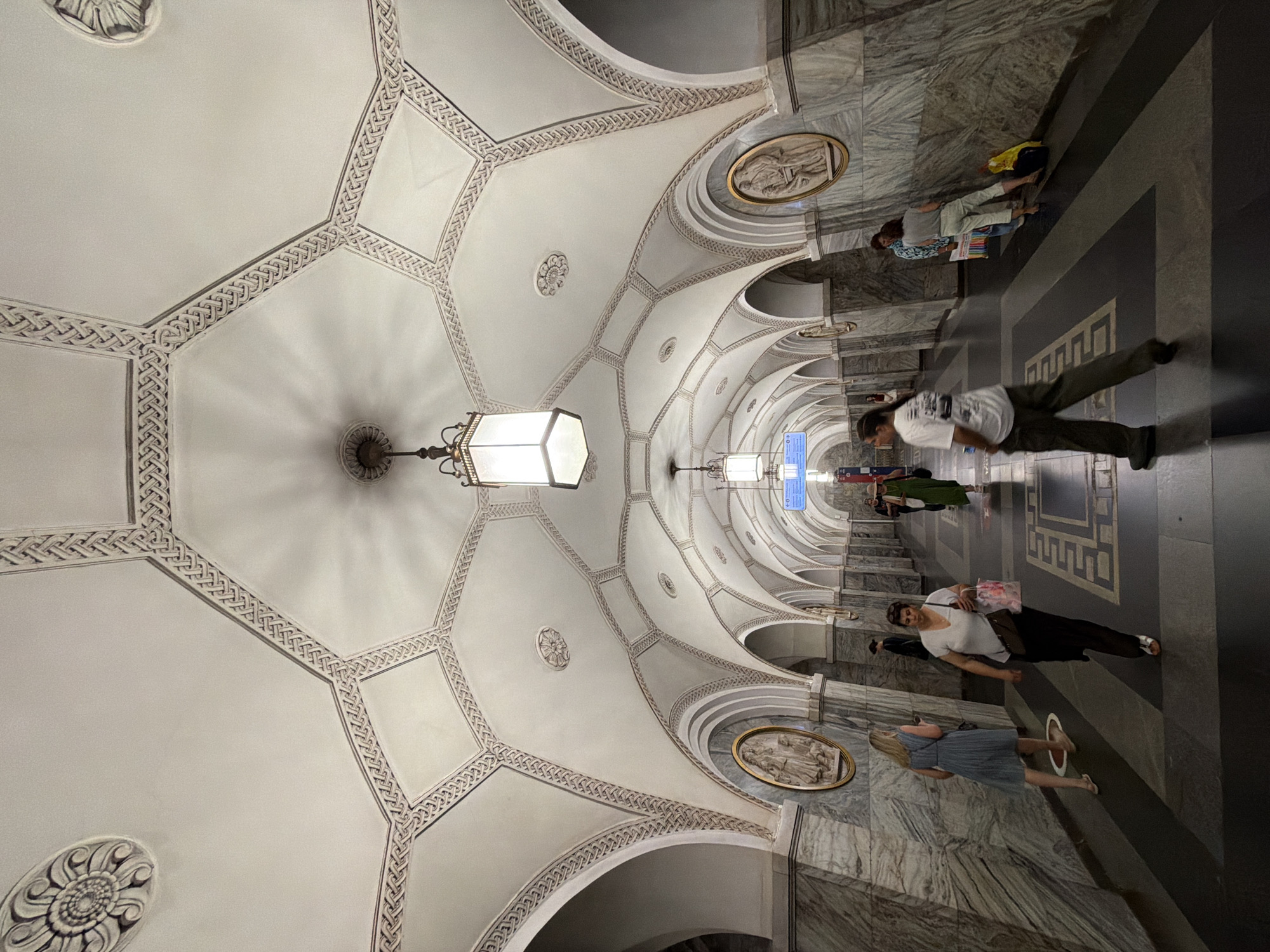 The image captures the ornate ceiling of a Moscow Metro station. The ceiling is characterized by its vaulted architecture, decorated with intricate geometric patterns and rosettes. Arched passages line the walls, which are clad in polished marble, creating a grand and somewhat somber atmosphere. Several people are seen walking through the station. In the foreground, a woman in a white t-shirt and black pants walks away from the camera, carrying shopping bags. To her left, a woman in a blue dress walks towards the camera, looking at her phone. Other individuals are visible further down the corridor, some interacting with each other or leaning against the wall. The station is illuminated by several rectangular pendant lights. The floor is a dark tiled surface with geometric patterns. This particular style of architecture and decoration is characteristic of many Moscow Metro stations, suggesting this is likely one of the historic and elaborately designed stations within the city.