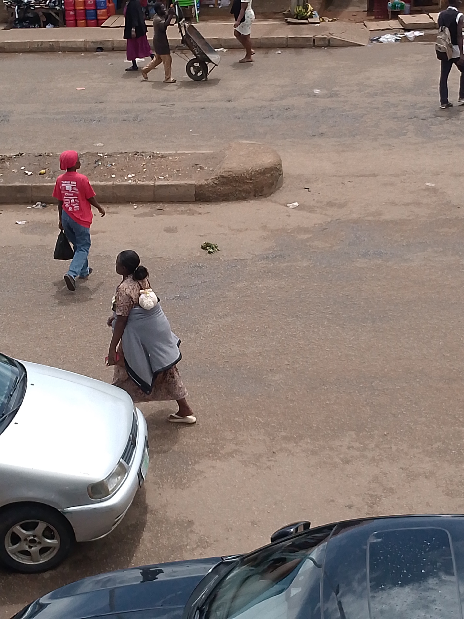 In Bukuru, Nigeria, a street scene unfolds with various individuals and vehicles. In the foreground, a silver car is partially visible, its front end angled towards the left. Behind it, the roof of a black car is seen. A woman, with her hair tied in a bun, carries a baby wrapped in a grey cloth across her back, walking towards the left of the frame. She is wearing a patterned dress and appears to be wearing sandals. To her left, a young boy wearing a red cap and a red t-shirt with "THANK YOU" printed on the back is walking away from the camera. He is carrying a black bag in his left hand and wearing blue jeans. Further in the background, other individuals are present, including a person pushing a wheelbarrow. The environment appears to be a dusty street with some debris scattered around. In the far background, market stalls with colorful containers are visible.