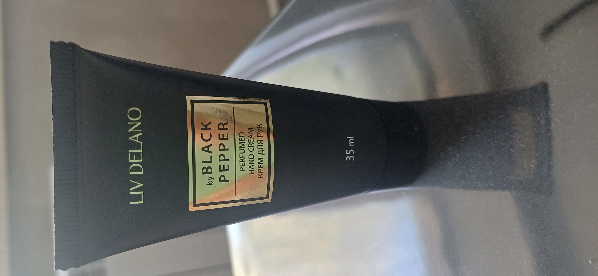 A black tube of hand cream with a gold label is shown against a gray background. The label reads "LIV DELANO" at the top, and "by BLACK PEPPER" below. Underneath that, it says "PERFUMED HAND CREAM" and "КРЕМ ДЛЯ РУК". The bottom of the tube shows "35 ml". There are some minor smudges on the tube and label. The product is placed on a reflective surface, casting a shadow. There is no indication of the location being Ekibastuz, Kazakhstan.