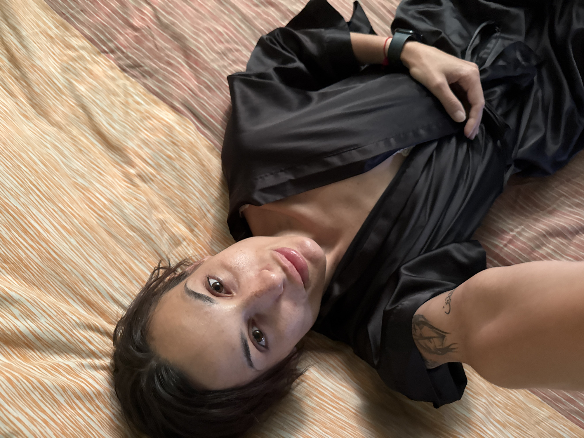 A woman is lying on a bed with orange and pink striped bedding. She is wearing a black satin robe. Her left arm is visible, with tattoos on her forearm. Her right hand is resting on her lap. She is looking at the camera with a slight smile. The background is a blurred orange and pink striped blanket. The overall scene appears to be a casual, indoor setting. The provided image does not contain any information or visual cues that would allow for the identification of the location as Pleven, Bulgaria.