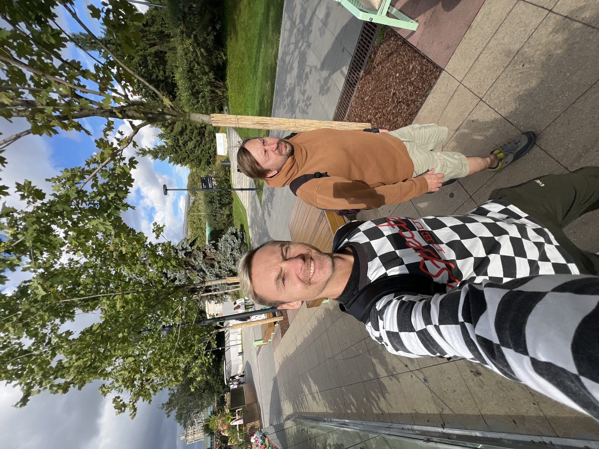 Two men are standing on a paved walkway in a park-like setting. The man in the foreground is taking a selfie and is wearing a black and white checkered long-sleeved shirt and a black backpack. He has blond hair and is smiling. The man behind him is wearing a brown hooded sweatshirt and light-colored shorts. He has brown hair and is looking to his left with his eyes partially closed. In the background, there are trees, benches, and other park amenities. The sky is partly cloudy. The man in the foreground has a logo on his shirt that reads "Special Duty". The location is identified as Moscow, Russia.