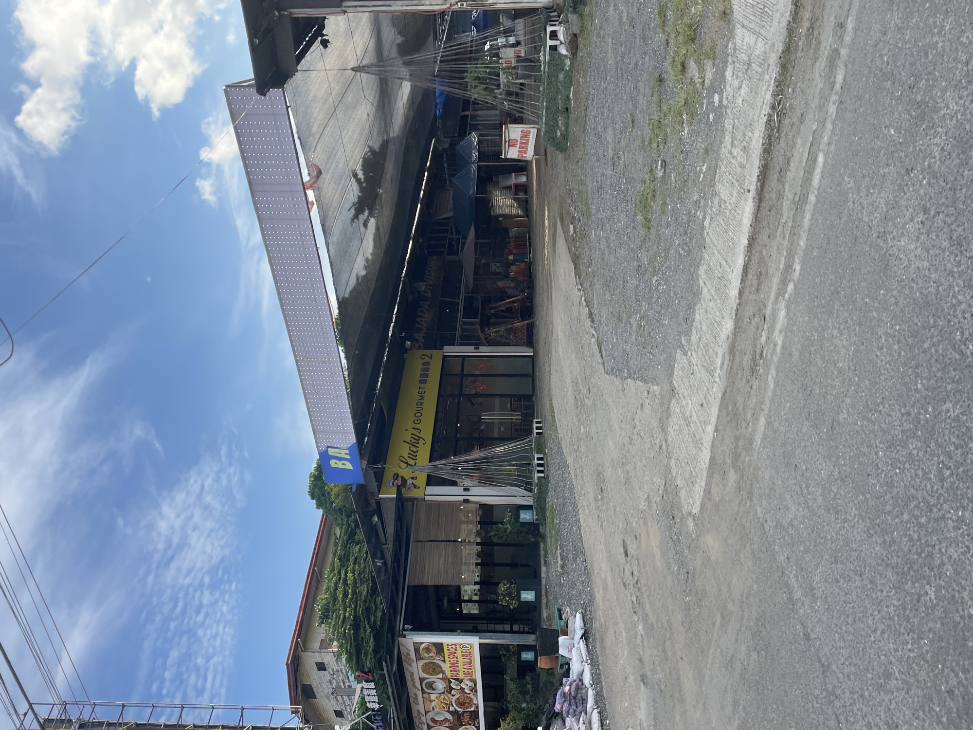 The image depicts the exterior of a commercial establishment in Davao City, Philippines, on a bright, partly cloudy day. The foreground shows a rough, unpaved road leading towards the building.
The building itself appears to be a single-story structure with a dark, slatted overhang providing shade. A prominent yellow sign with red lettering reads "Lucky's GOURMET" followed by some text in what appears to be Chinese characters. To the left of this sign, a smaller sign displays images of food and text indicating "PARKING SPACES ARE AVAILABLE".
Behind the main building, a large, white, rectangular structure with many small lights is visible, possibly a large LED display or signage. A black mesh net is stretched over a section of the area in front of the building. Various items are displayed for sale or in storage under the covered area, including what appear to be umbrellas, wooden furniture, and stacks of goods. A "NO PARKING" sign is placed on the right side of the scene.
In the background, a multi-story building with a red roof and white brick facade is visible, surrounded by lush green trees. Overhead, utility wires stretch across the frame, and a tall metal structure, possibly a utility pole or scaffolding, is present on the far left. The overall impression is of a commercial street or market area.