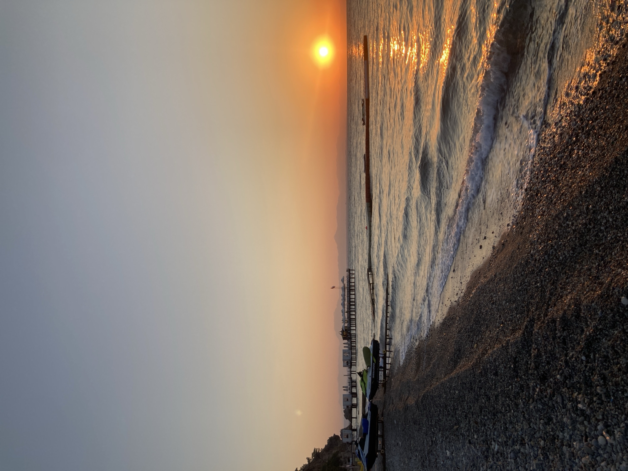 This image depicts a tranquil sunset scene at a pebble beach in Kemer, Turkey. The sun, a vibrant orange orb, is low on the horizon, casting a warm golden glow across the calm sea. Gentle waves lap against the shore, reflecting the sunlight.
In the foreground, a dark pebble beach is visible, with two jet skis parked on stands. Further out in the water, several piers or jetties extend from the shore. Some of these structures have small buildings or cabanas on them, and white umbrellas are arranged neatly. In the distance, silhouetted mountains form the backdrop to the sunset.
The sky transitions from a pale blue at the top to a soft orange near the horizon, indicating the twilight hour. The overall atmosphere is peaceful and serene, capturing the beauty of a sunset on the Mediterranean coast. There are no people visible in the image.