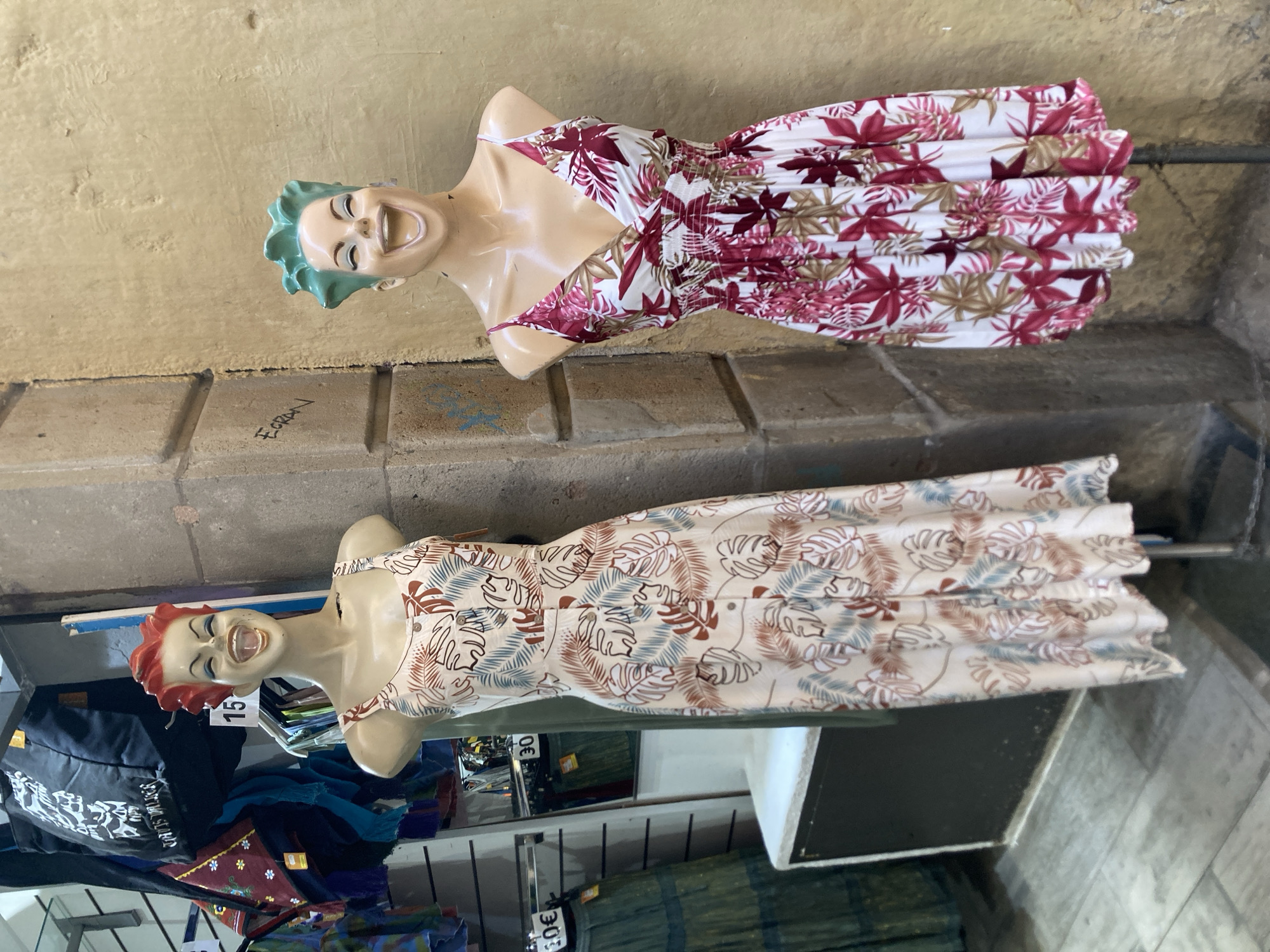 The image shows two mannequins wearing dresses with floral patterns, displayed in what appears to be an outdoor market stall. The mannequin in the foreground has red hair and is wearing a light-colored dress with brown and blue leaf designs. The mannequin in the background has green hair and is wearing a white dress with pink and burgundy floral patterns. Both mannequins have exaggerated, smiling faces. The background consists of a brick wall and a concrete floor. There are bags and clothing racks visible in the lower part of the image, suggesting a retail environment. The lighting suggests it is daytime. The image is oriented vertically, with the subjects appearing to be upright from their perspective. Visible text includes "15" and "30" which could be price tags. There is also some graffiti on the wall. The overall scene depicts a colorful display of clothing, likely in a busy marketplace.