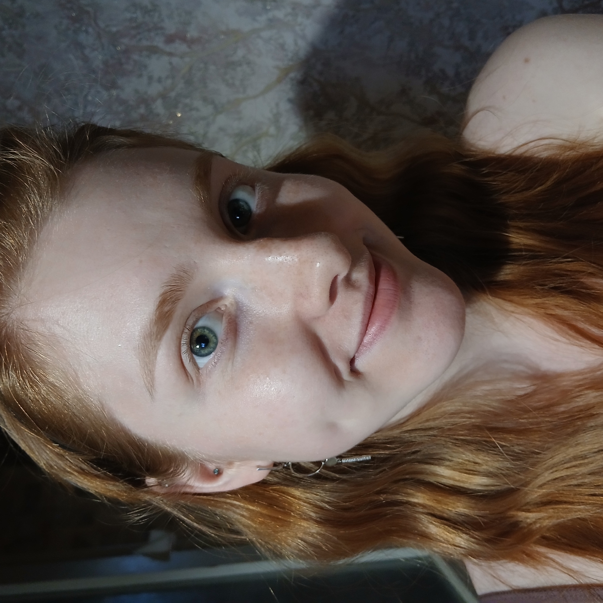 This close-up portrait features a young woman with long, wavy, reddish-brown hair, appearing to lie down as she looks upward towards the camera. The image is angled from above, with her head positioned in the bottom left, and her hair flowing diagonally across the frame to the upper right. She has fair skin, a scattering of light freckles across her nose, and bright, light-colored eyes, which appear to be greenish-blue. Her lips are slightly parted in a subtle, natural smile, conveying a calm and content expression.
On her visible left ear, she wears a small stud piercing in her tragus and a delicate dangle earring. Her skin shows a natural sheen on her forehead and cheeks. The background is indistinct, consisting of a mottled, textured surface in muted tones of grey and blue, with some shimmering highlights. A portion of her bare shoulder or upper chest is visible in the upper right. The lighting is soft and even, suggesting an indoor setting. The image captures a moment of serene contemplation or casual posing. No text is visible within the image. While the provided context suggests the location is Yekaterinburg, Russia, the visual details within the photograph do not offer specific cues to this geographic location.