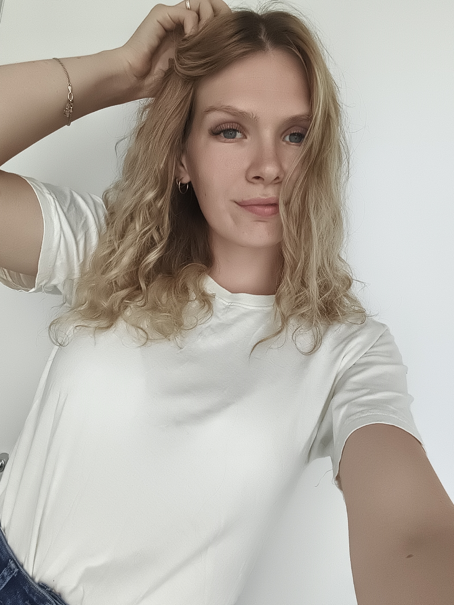 A young Caucasian woman with blonde, wavy hair and blue eyes is shown from the chest up, facing the camera. She is wearing a plain white t-shirt and has her right hand raised, touching her hair. A delicate gold bracelet is visible on her left wrist, and she is wearing a small hoop earring in her left ear. The background is a plain white wall. There is no discernible indication of the city of Izhevsk, Russia, in the image.