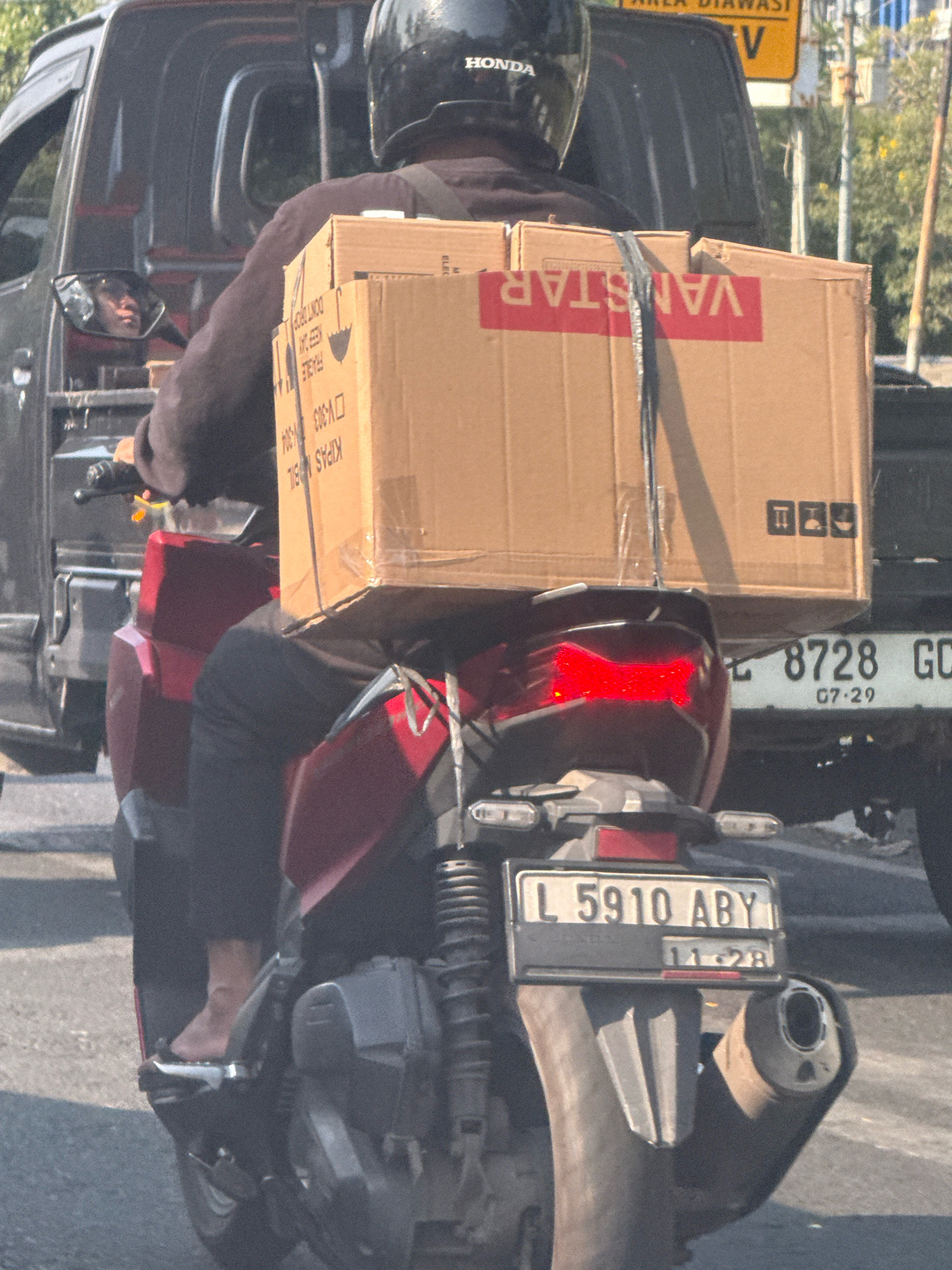 The image shows a person riding a dark red scooter on a street in Surabaya, Indonesia. The rider is wearing a black helmet with "HONDA" branding and a dark jacket. They are carrying two large cardboard boxes strapped to the back of the scooter. The boxes have red and white branding that appears to say "VANSTAR." The scooter has a license plate that reads "L 5910 ABY" and a smaller sticker below it indicating "11-28". In the background, a dark colored pickup truck is visible to the left of the scooter. The street is paved, and there are some trees and buildings in the distance. The overall lighting suggests it is daytime.