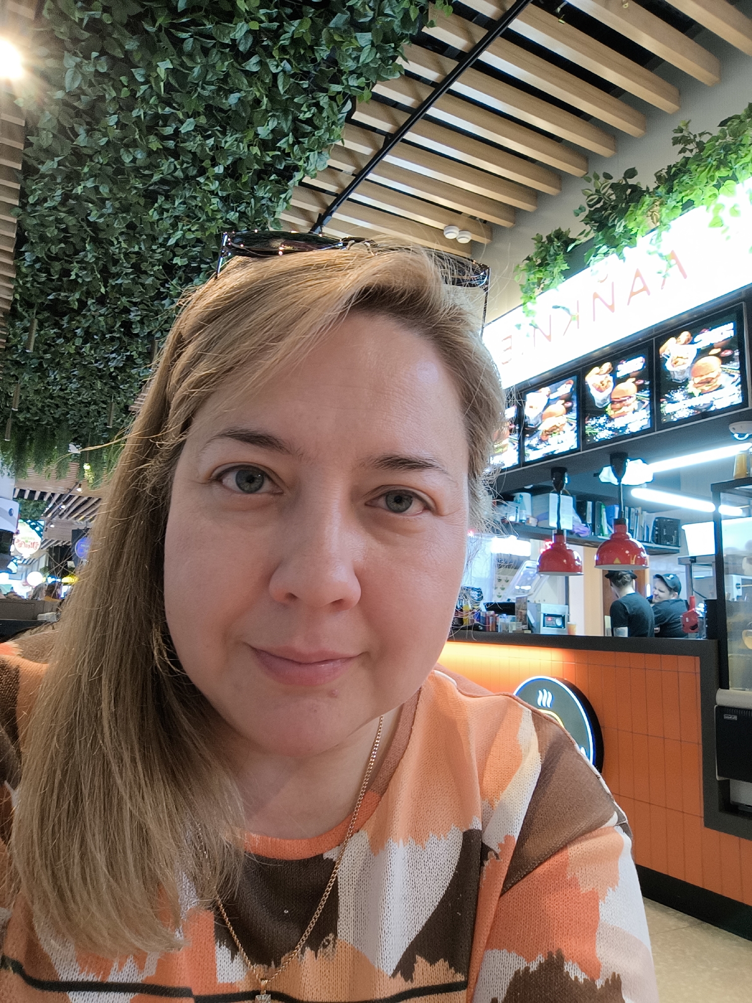 A woman is taking a selfie in a restaurant. She has light-colored hair and is wearing a patterned sweater. On top of her head rests a pair of sunglasses. In the background, there is a food service counter with menu boards displaying images of hamburgers. Two workers stand behind the counter. The restaurant's decor includes artificial greenery and a wooden slat ceiling. The sign above the menu displays the word 'ENKNAKE' in reverse, which suggests this is a location of the restaurant in Moscow, Russia.
