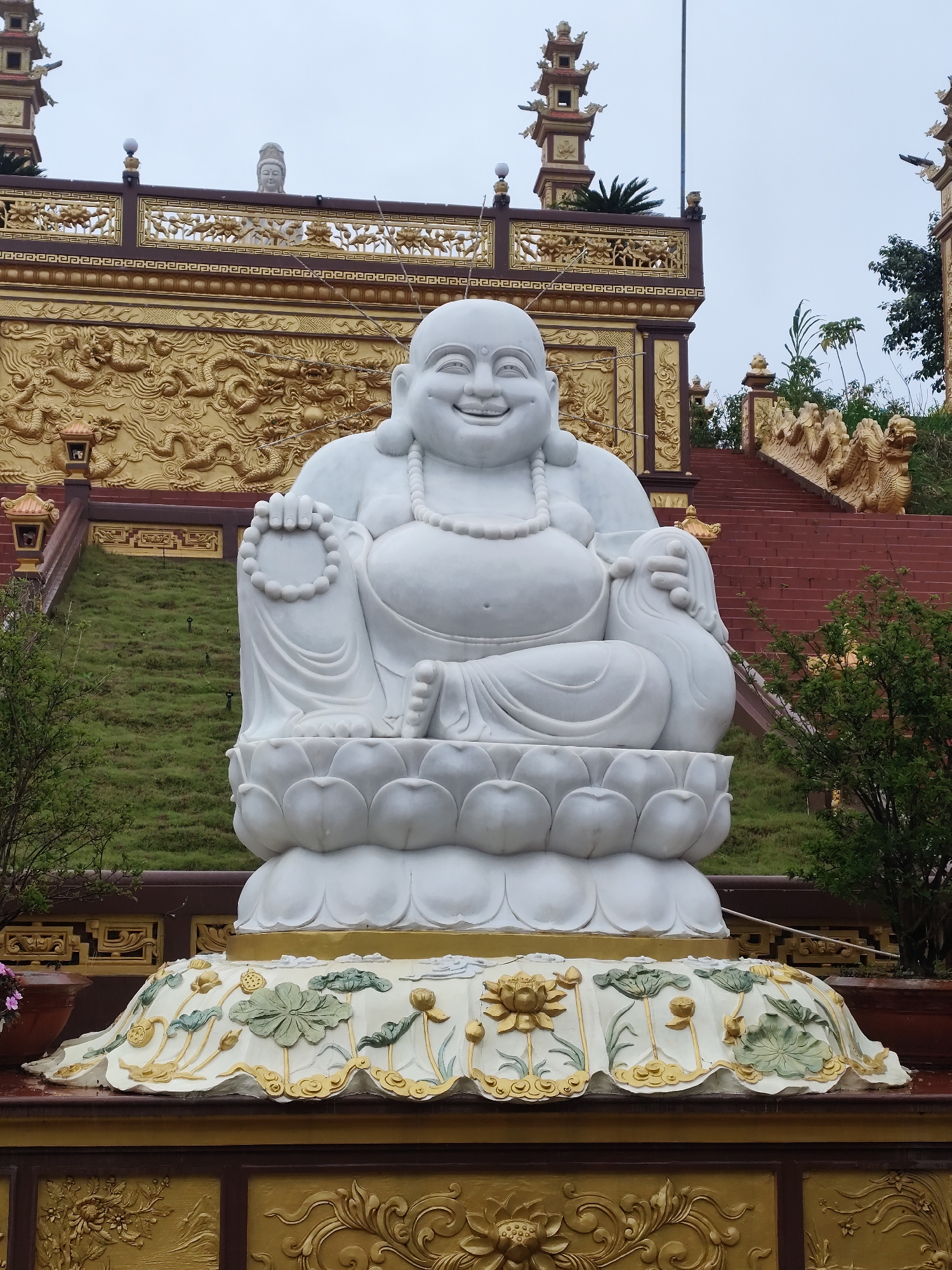 A large white marble statue of a laughing Buddha is prominently displayed outdoors, seated on a lotus flower base. The statue has a serene expression, a full belly, and is adorned with a beaded necklace. The base of the statue is intricately decorated with relief carvings of lotus flowers and leaves in green and gold. Behind the Buddha statue, a red staircase with dragon-shaped handrails leads upwards towards a golden wall adorned with elaborate dragon reliefs. Further behind the wall, traditional Asian pagodas with tiered roofs are visible against an overcast sky. Lush green grass covers a sloping hill to the left of the staircase. The scene appears to be within a temple complex. The statue is located at the Lạc Dương Temple in the city of Lạc Dương, Vietnam.