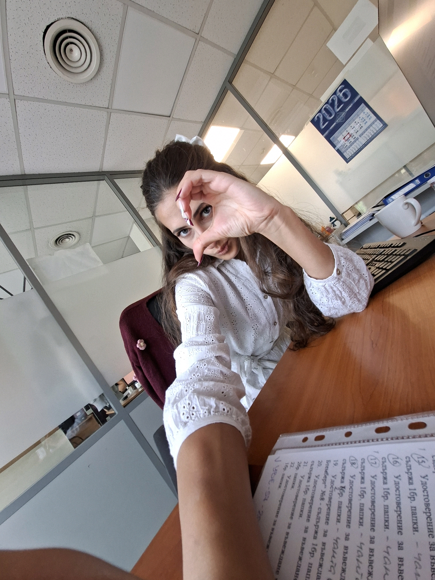 A young woman with long dark hair, wearing a white eyelet blouse, sits at a desk in an office. She is holding her hands up, forming a heart shape over her face as she looks directly at the camera. Her nails are painted with a red and white design. The office environment is visible, including a ceiling vent, glass partitions, a keyboard, a white mug, and a calendar on the wall showing the year 2026. A binder with papers is on the desk in the foreground.