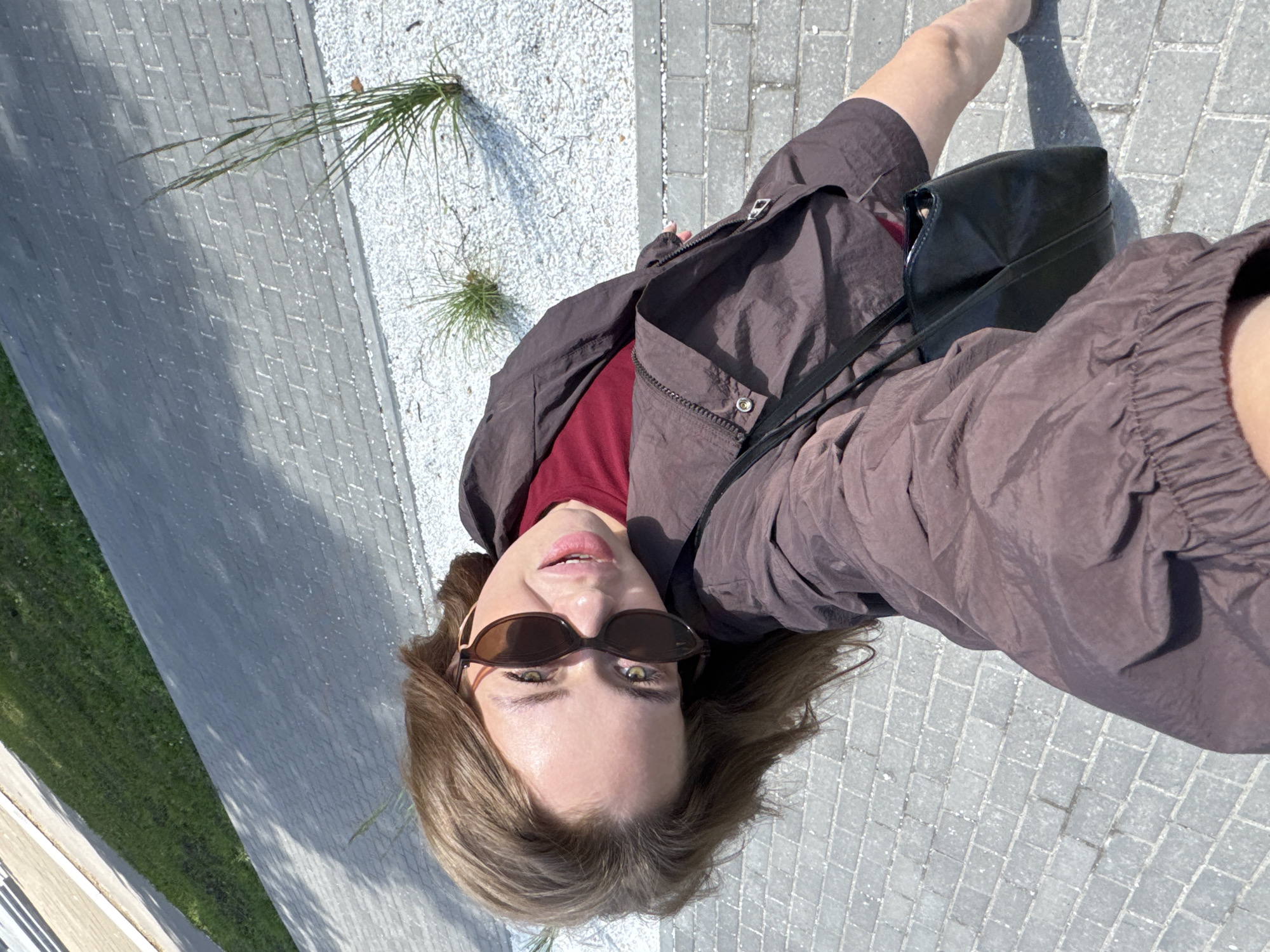 A young woman with light brown hair, wearing sunglasses, a dark brown jacket over a maroon t-shirt, and holding a black purse, is taking a selfie outdoors. She is standing on a paved walkway made of rectangular grey bricks. To her right, there is a section of white gravel with sparse green grass growing in it. Beyond the gravel is a strip of green grass, and further in the background, another paved area. The sun is casting shadows on the walkway. The woman's pose and the perspective suggest she is looking down at the camera while holding it at arm's length. The scene appears to be in a public outdoor space, possibly a park or a pedestrian area within the city of Chaikovsky, Russia.