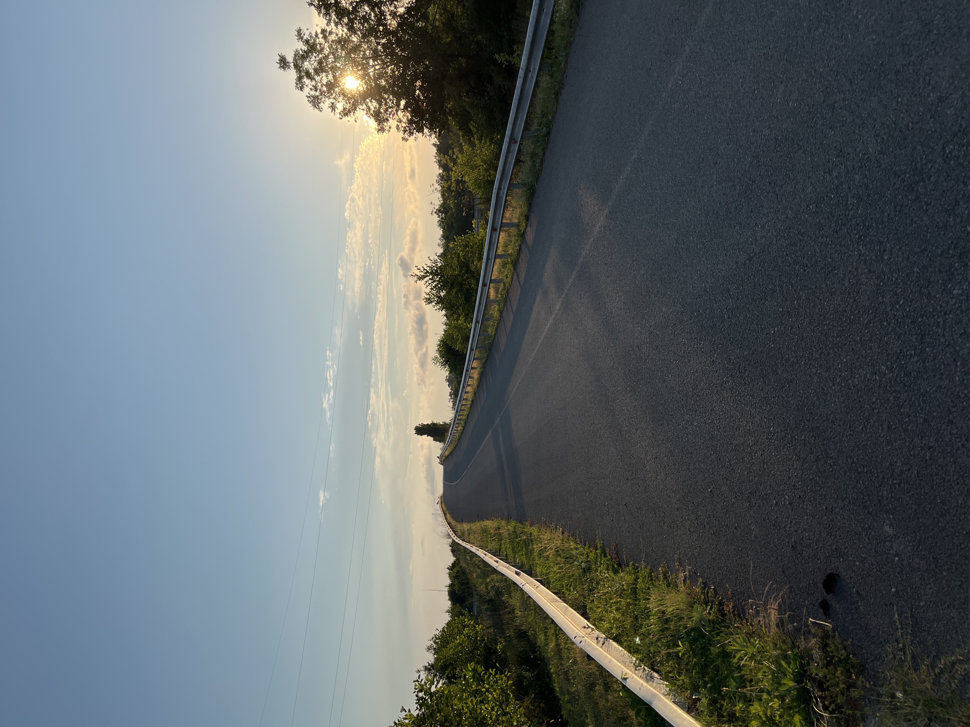 This is a low-angle, vertical shot of a paved road in Ureki, Georgia, leading uphill towards a bright sunset. The road has a metal guardrail on the left side, separating it from a grassy embankment with scattered trees. On the right side, the guardrail is also visible, bordering a line of dense green trees. The sun is setting behind these trees on the right, casting a warm glow and dappling sunlight onto the road and surrounding foliage. Power lines are strung across the clear blue sky, with a few wispy clouds near the horizon. The overall atmosphere is serene and natural, with the road appearing empty of people or vehicles.