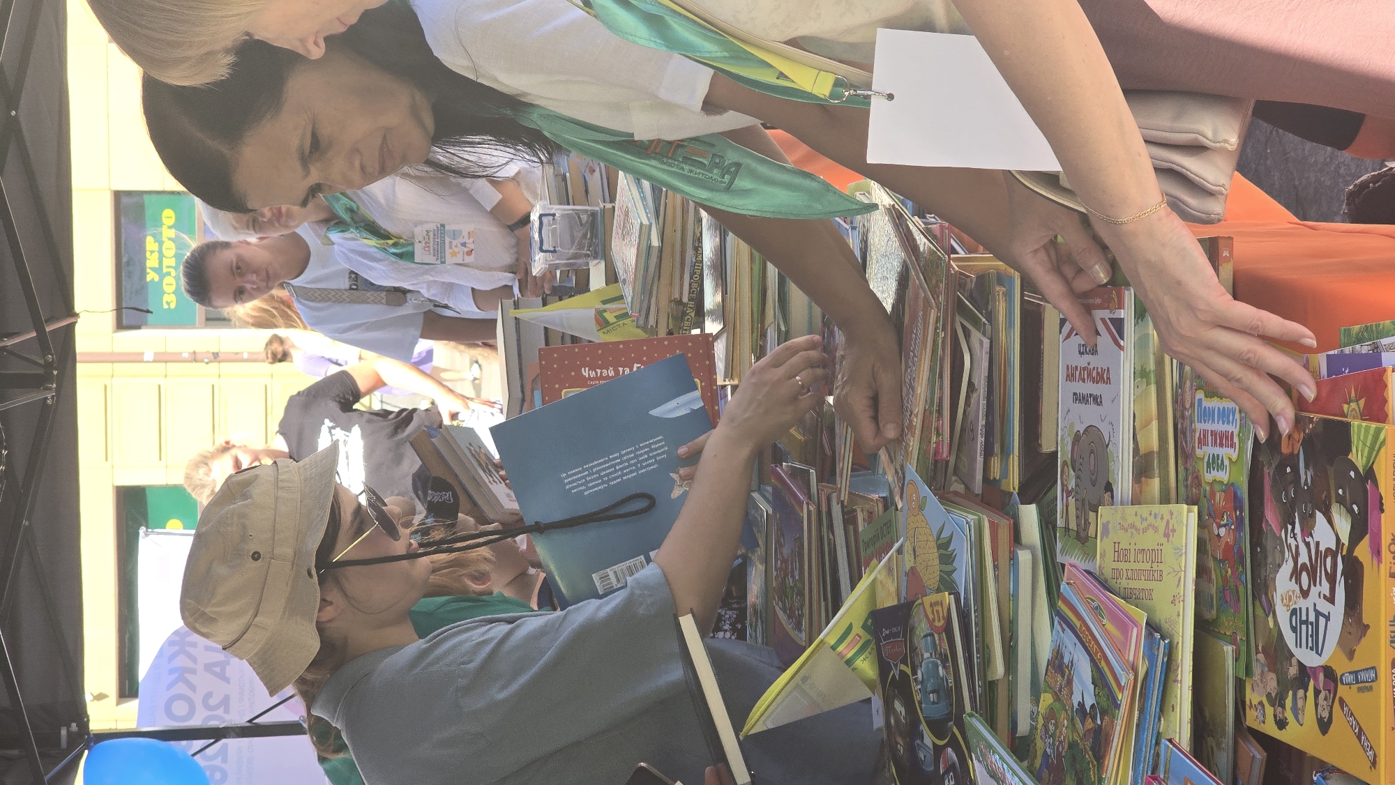 The image captures an outdoor book sale event in Zhytomyr, Ukraine. In the foreground, a woman wearing a light-colored bucket hat and sunglasses is examining books. She is dressed in a blue top and is holding a notebook in her left hand. In the middle ground, a woman with dark hair is reaching towards a stack of books, and another woman with blonde hair is partially visible. Several people are browsing a large table laden with books of various sizes and colors. Behind the table, a tent provides shade, and a building with yellow signage is visible in the background. The overall atmosphere suggests a vibrant community event focused on literature.